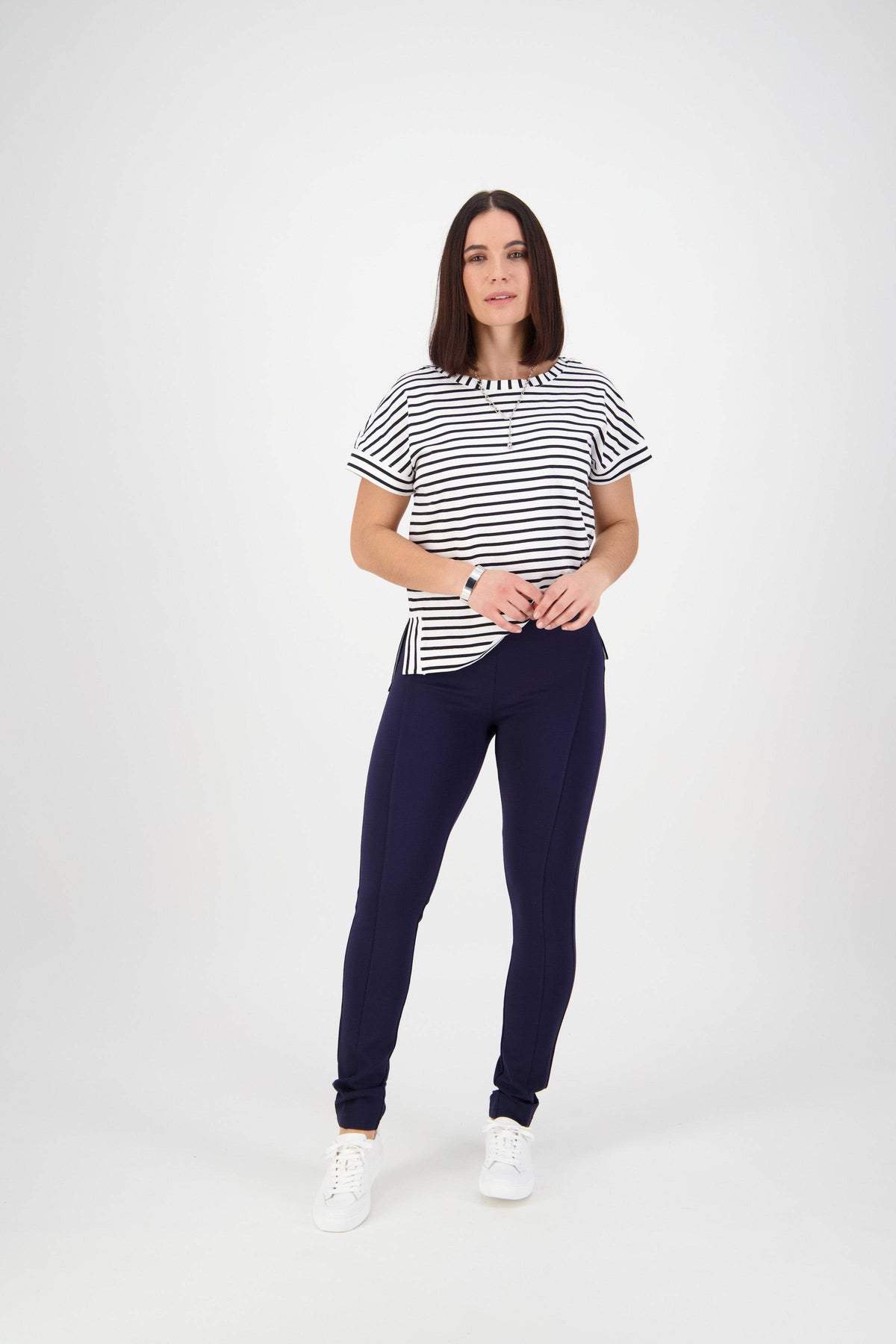 A woman with straight brown hair stands against a white background, wearing a black and white striped tee, Vassalli Toll Pant - Navy in stretch fabric, and white sneakers. She faces forward with her hands on her hips.