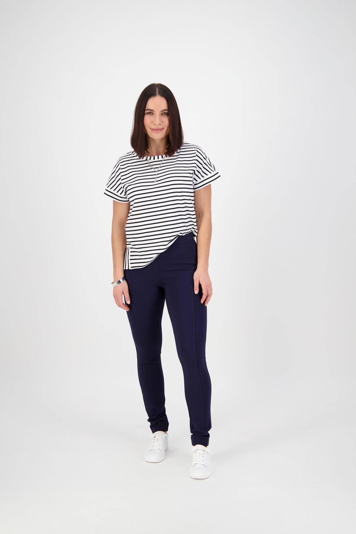 A woman with straight, shoulder-length brown hair faces the camera, wearing a black-and-white striped t-shirt, Vassalli Toll Pant - Navy skinny pull-on pants in stretch fabric, and white sneakers against a plain white background.