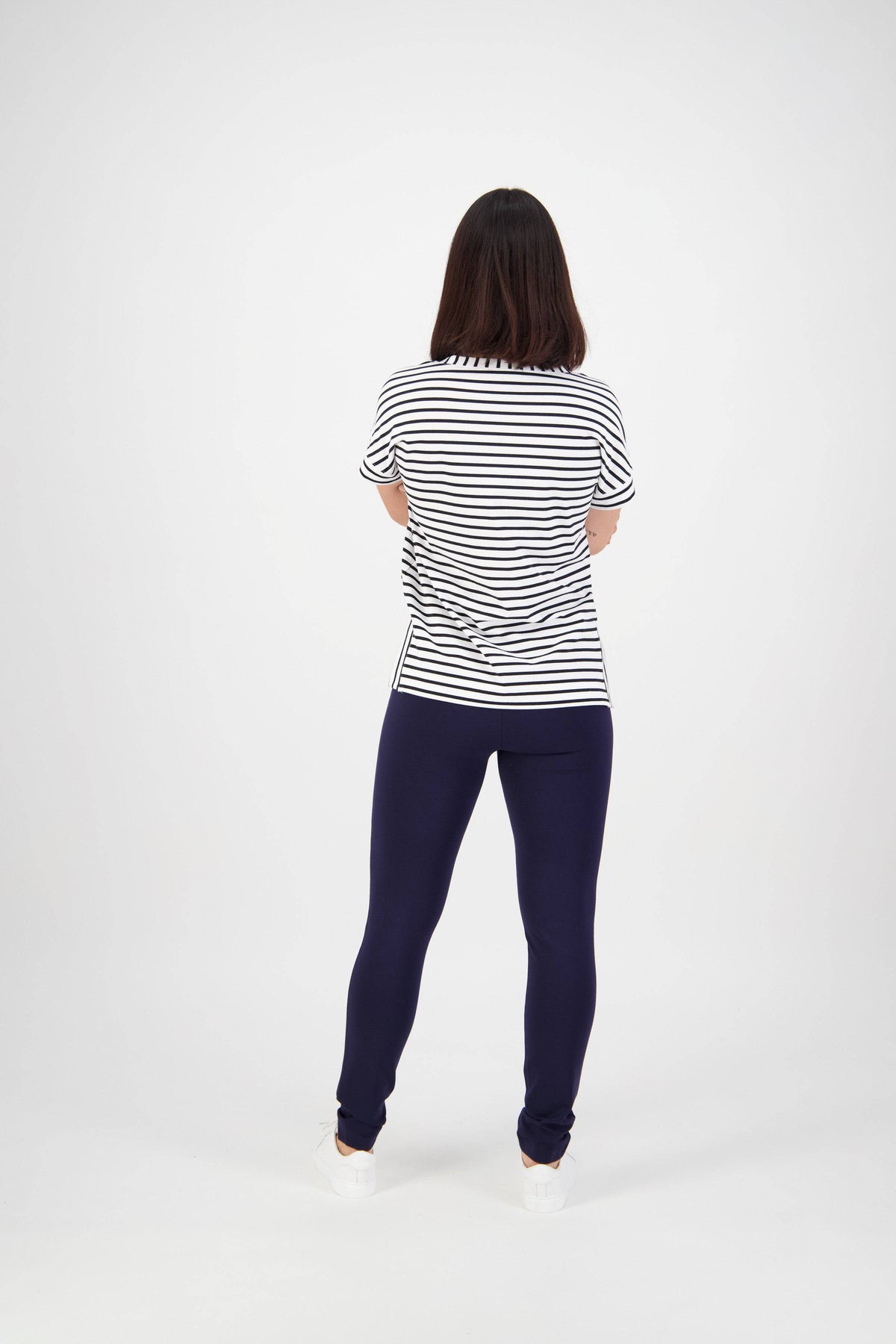 A woman faces away from the camera, wearing a short-sleeved black and white striped shirt, Vassalli Toll Pant - Navy, and white shoes against a plain white background.