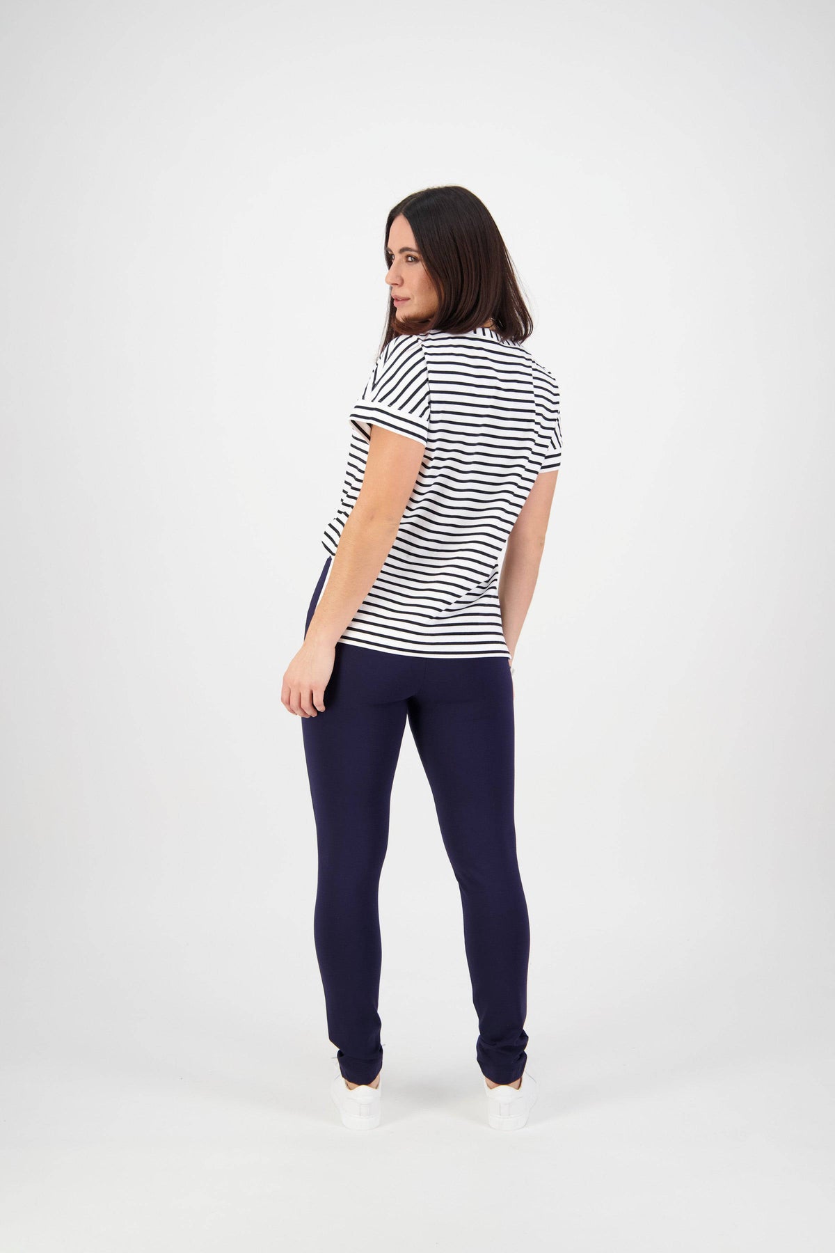 A woman with shoulder-length dark hair stands slightly turned, wearing a short-sleeved white shirt with black stripes and Vassalli Toll Pant - Navy, made from stretch fabric, against a plain white background.