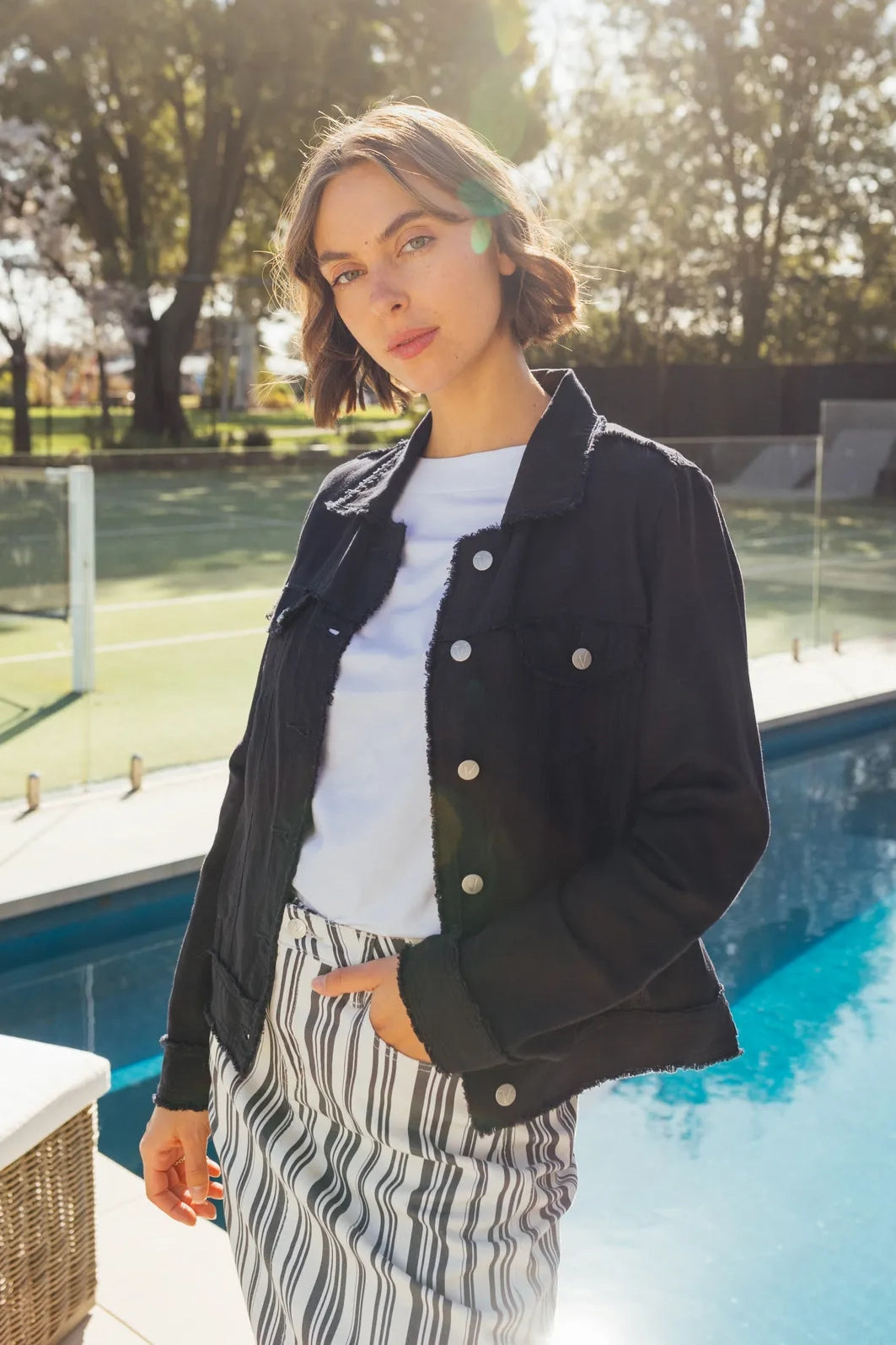 A woman with short hair wears a navy Vassalli Aura Jacket over a white top and striped pants, standing by a pool with sunlight shining behind her and trees in the background.