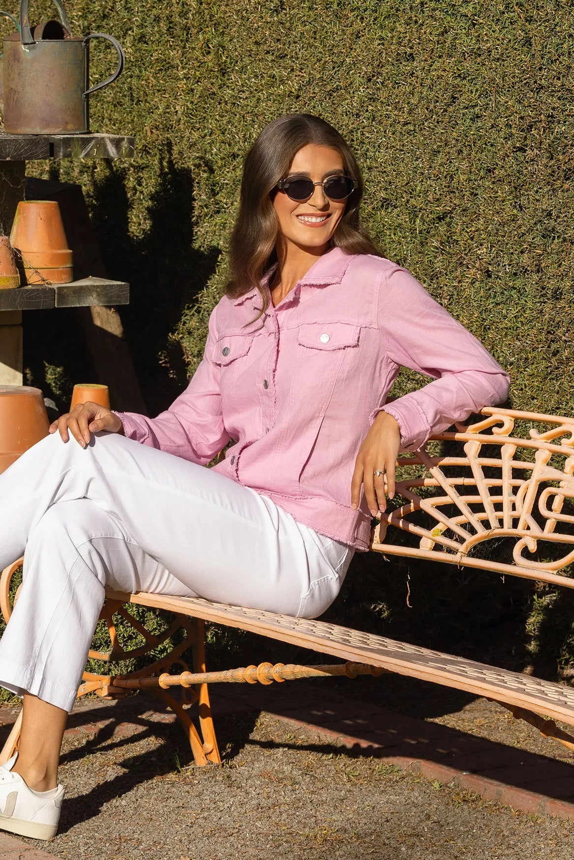 A woman wearing sunglasses, the Vassalli Aura Jacket in Taffy, and white pants sits smiling on a decorative outdoor bench with clay pots and a green hedge in the background.