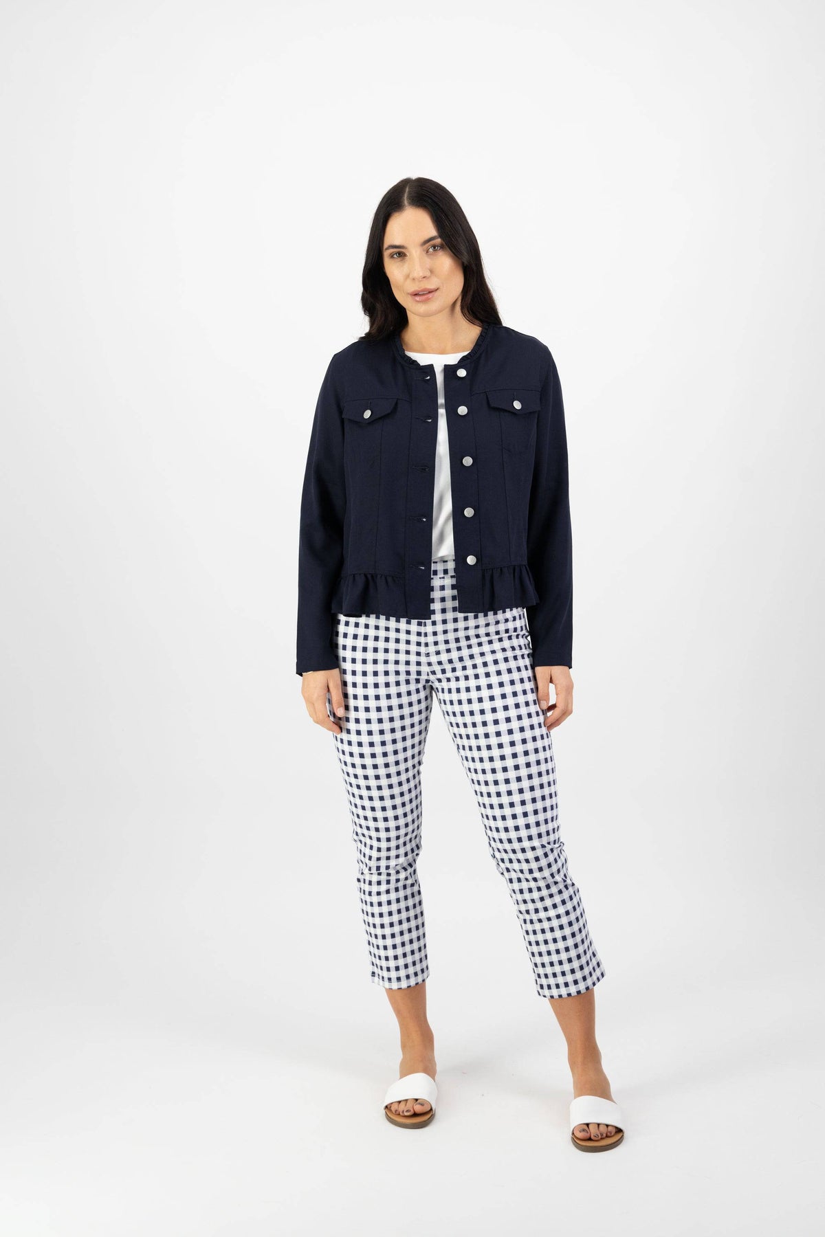 A woman with long dark hair and a neutral expression wears the Vassalli Sawyer Jacket - Ink over a white top, paired with blue and white checkered pants and white slide sandals, standing against a plain white background.