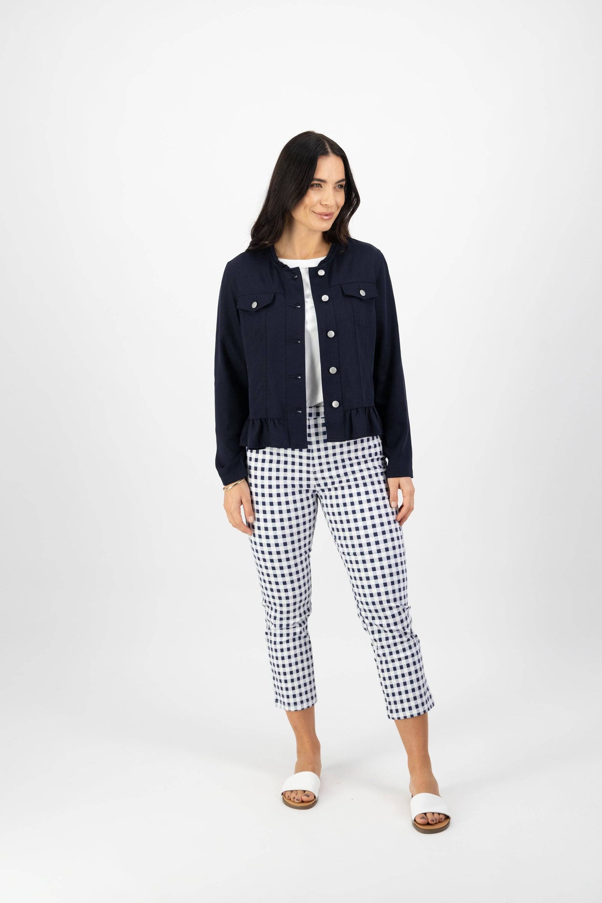 A woman with long dark hair and a relaxed expression stands against a white background, wearing the Vassalli Sawyer Jacket in Ink over a white top and blue-and-white checkered pants, paired with white slide sandals.