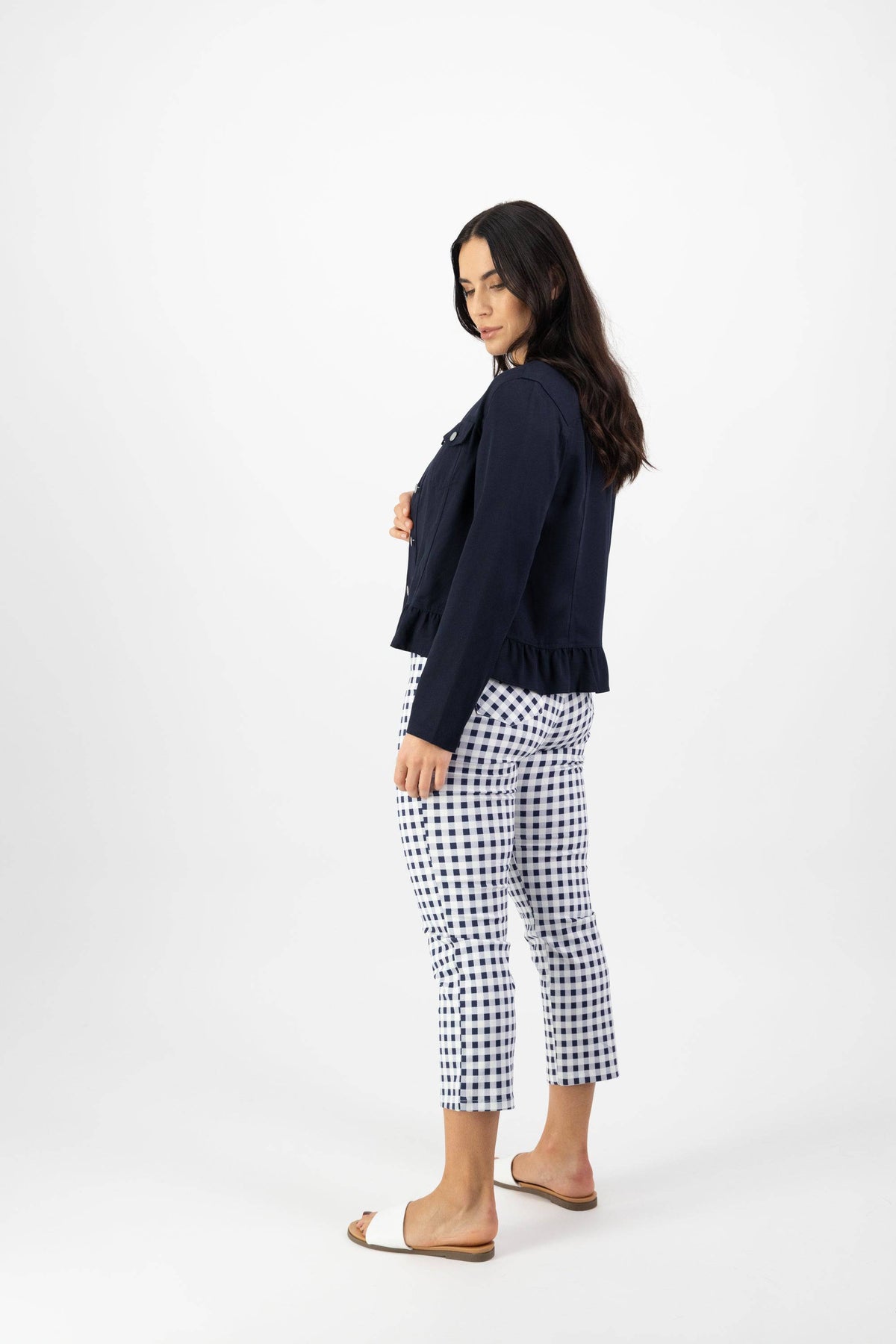 A woman with long dark hair stands sideways against a white background, wearing the Vassalli Sawyer Jacket in Ink, white and navy checkered cropped pants, and tan slide sandals.