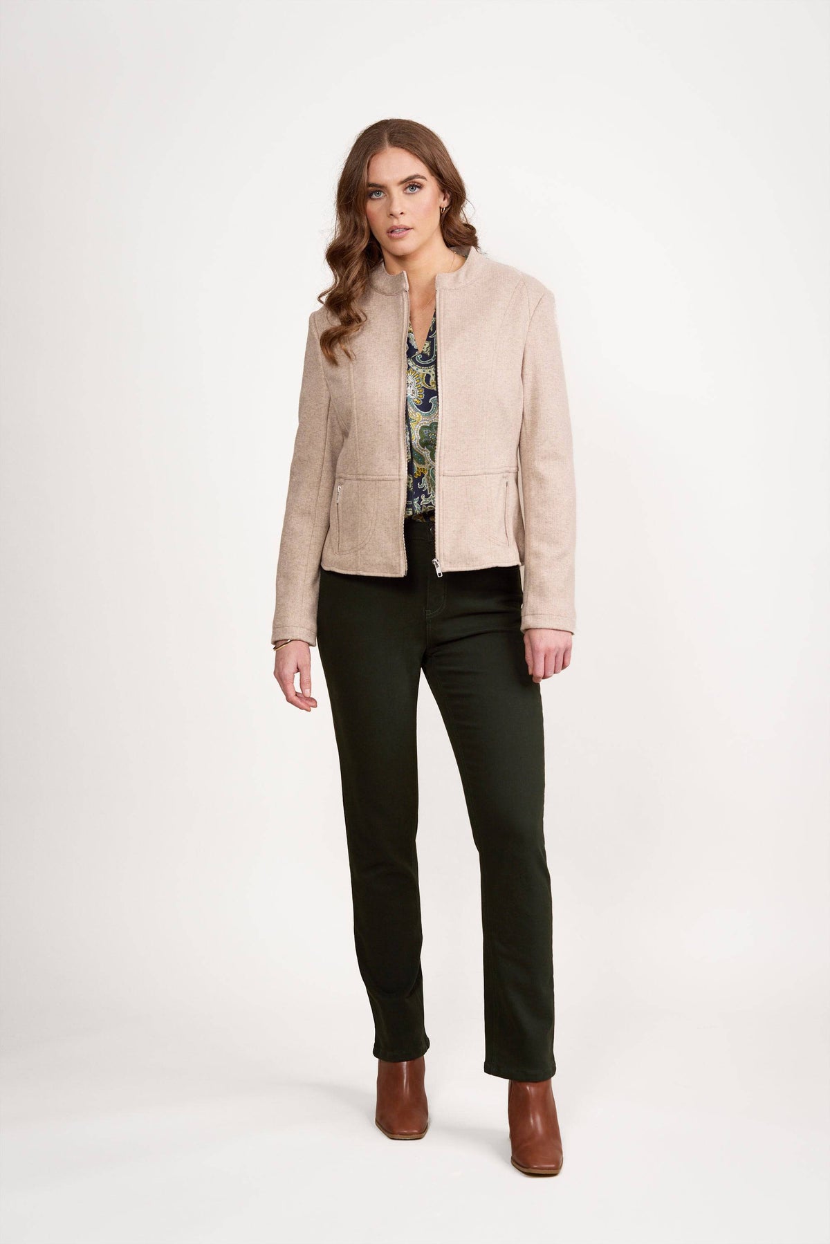 A woman with long wavy hair wears the Vassalli Zip Up Jacket in Dune, a patterned blouse, dark pants, and brown ankle boots, standing against a plain white background.