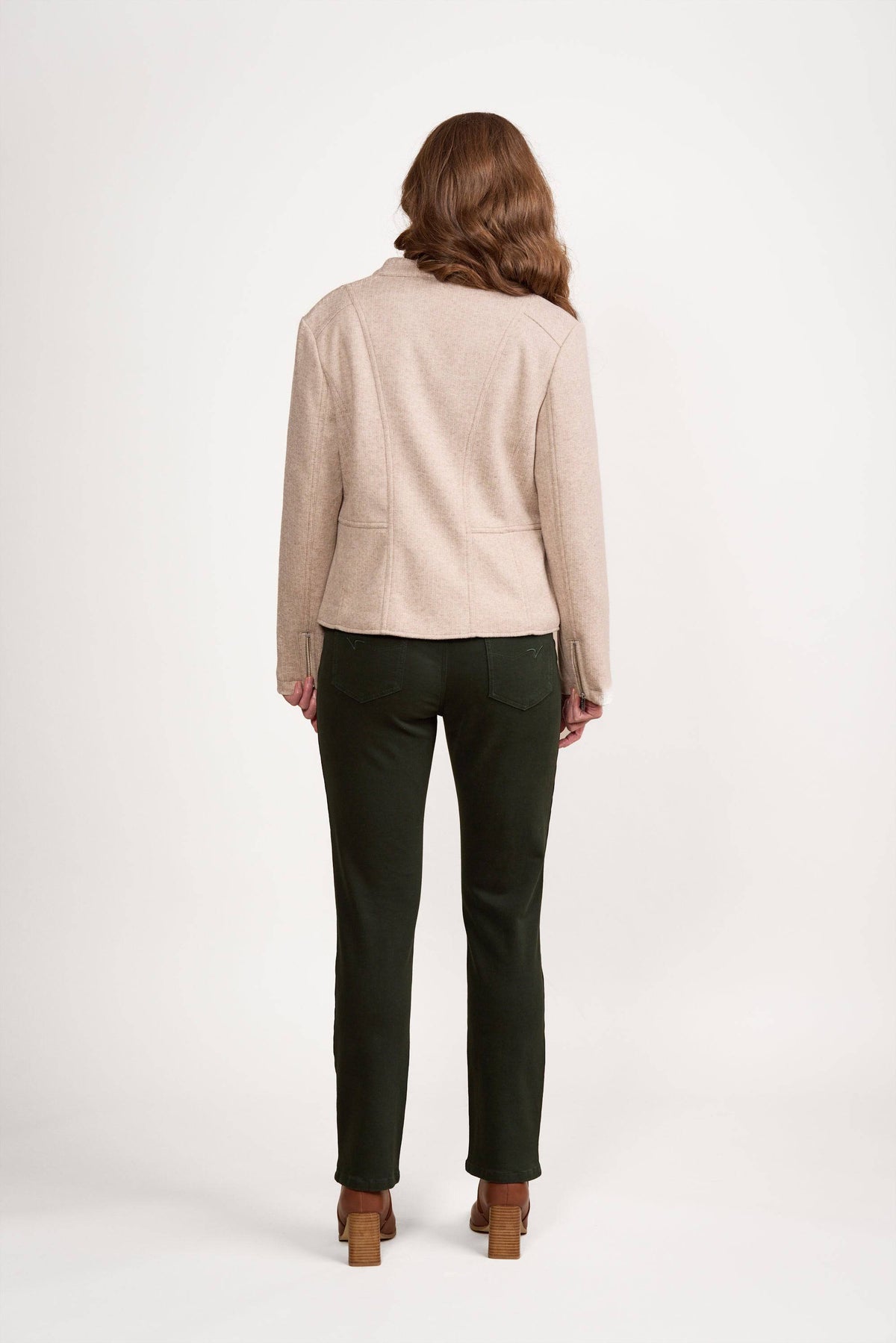 A woman stands facing away against a plain white background, wearing the Zip Up Jacket - Dune by Vassalli, along with dark green pants and brown heeled shoes.