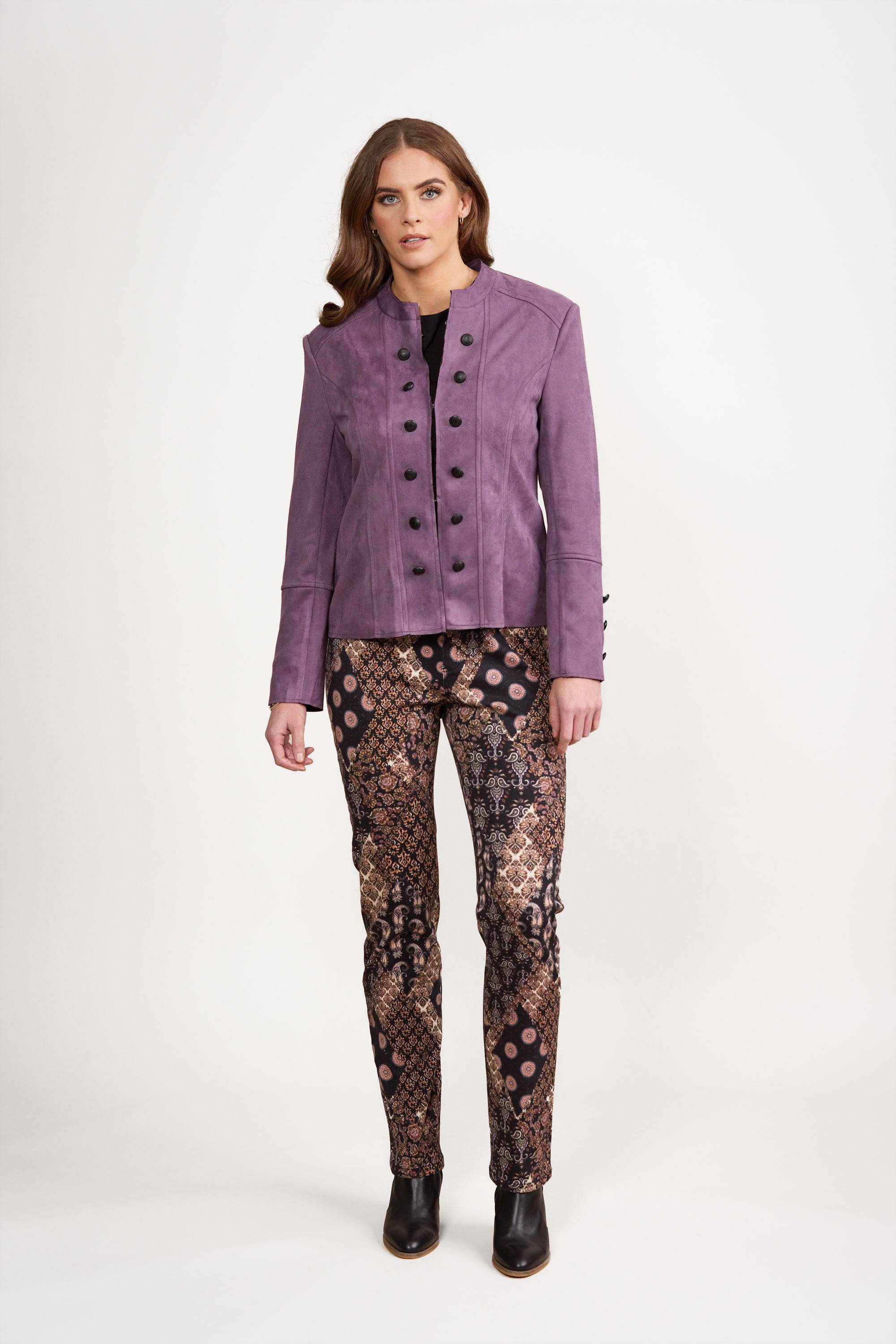 A woman stands against a plain white background wearing the Vassalli Military Jacket in Grape, paired with patterned pants and black shoes.