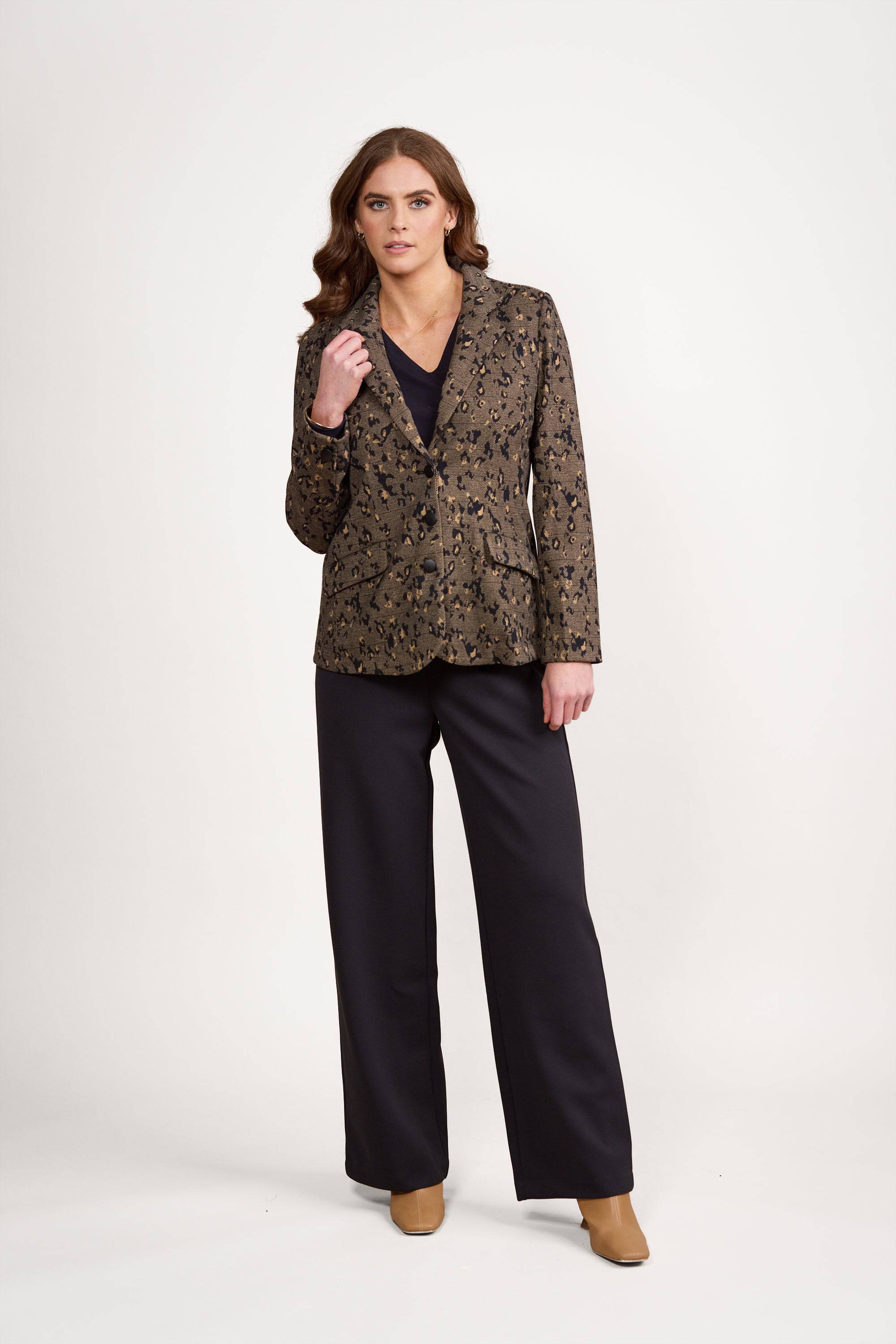 A woman with long brown hair, dressed in the Vassalli Knit Blazer - Globe over a black top and wide-leg black trousers, paired with tan shoes, stands against a plain white background.