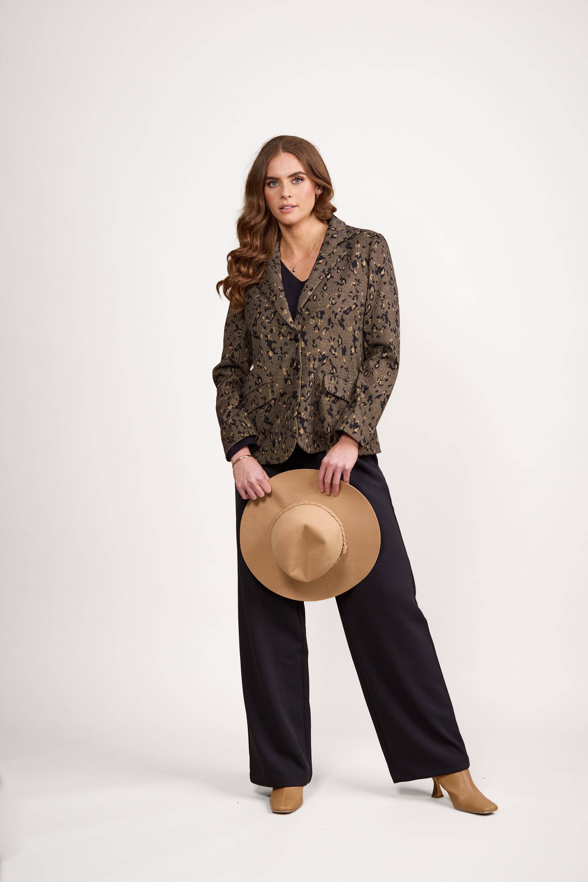 A woman with long brown hair stands against a white background, wearing the Vassalli Knit Blazer - Globe over black pants and tan boots. She holds a tan hat in front of her with both hands and looks directly at the camera.