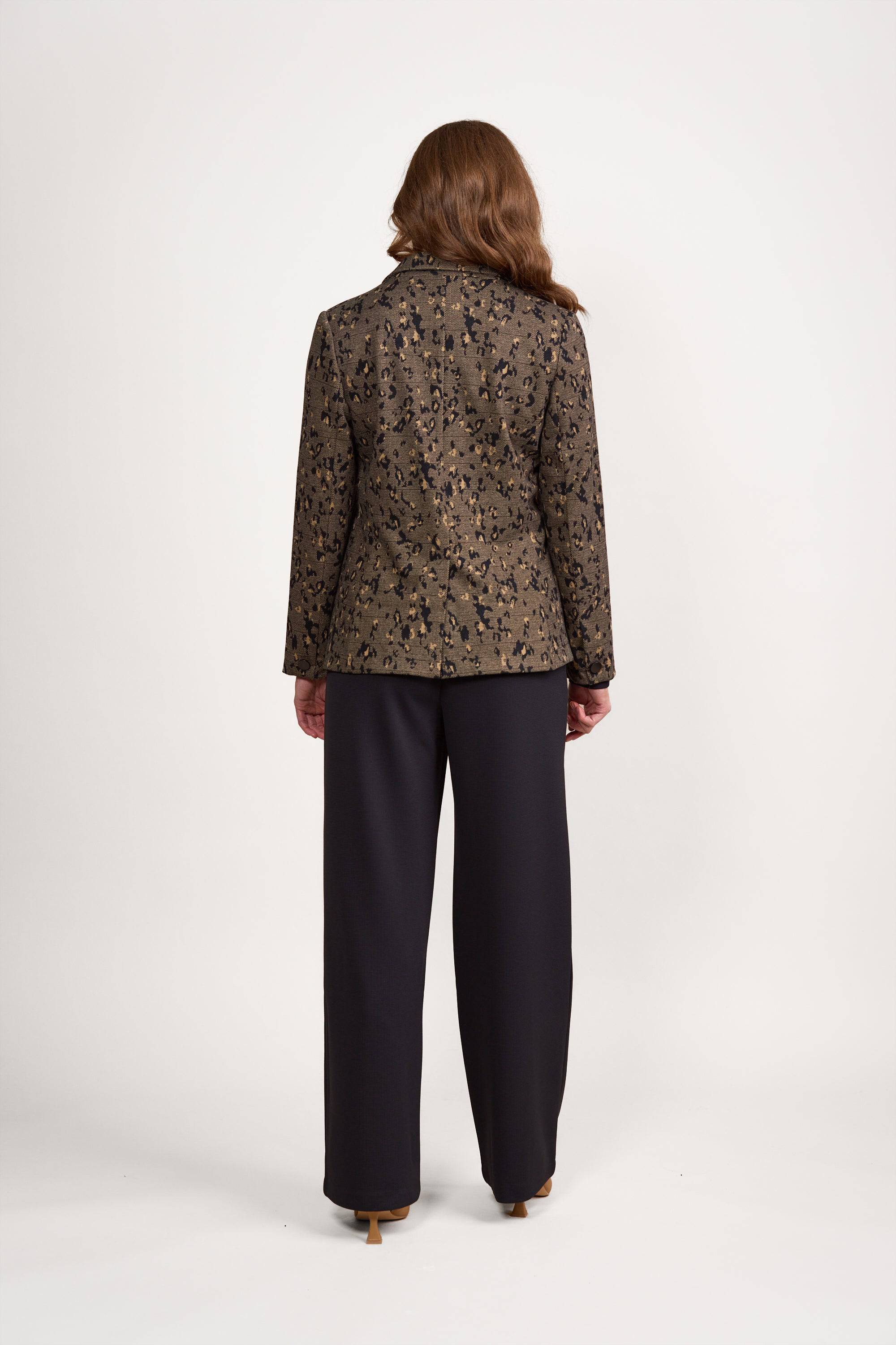 A woman stands facing away, wearing the Vassalli Knit Blazer - Globe featuring a modern pattern in brown, paired with black wide-leg pants. Her wavy hair cascades down her back as she poses against a plain white background.