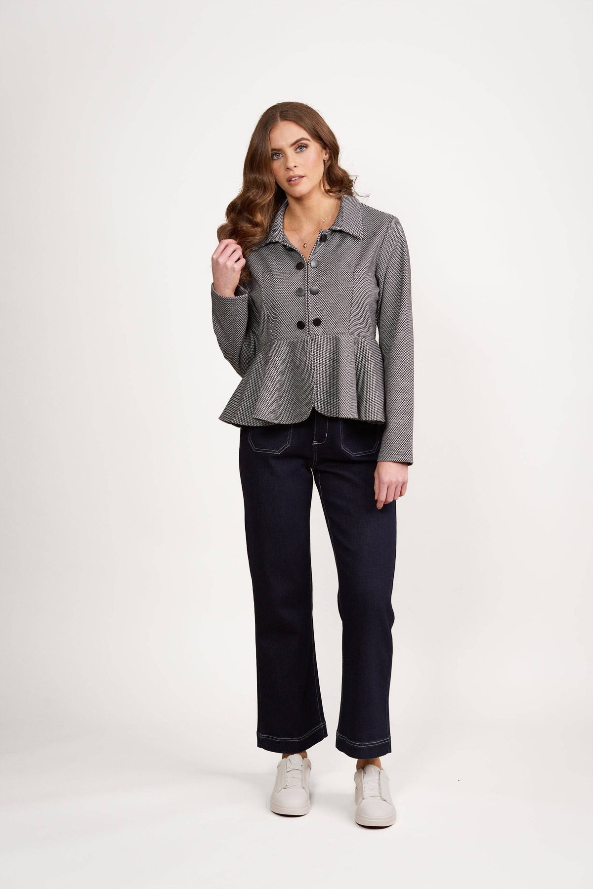 A woman with wavy brown hair wears the Vassalli Knit Jacket - Xander in gray, paired with dark wide-leg jeans and white shoes, standing against a plain white background and looking at the camera with a neutral expression.