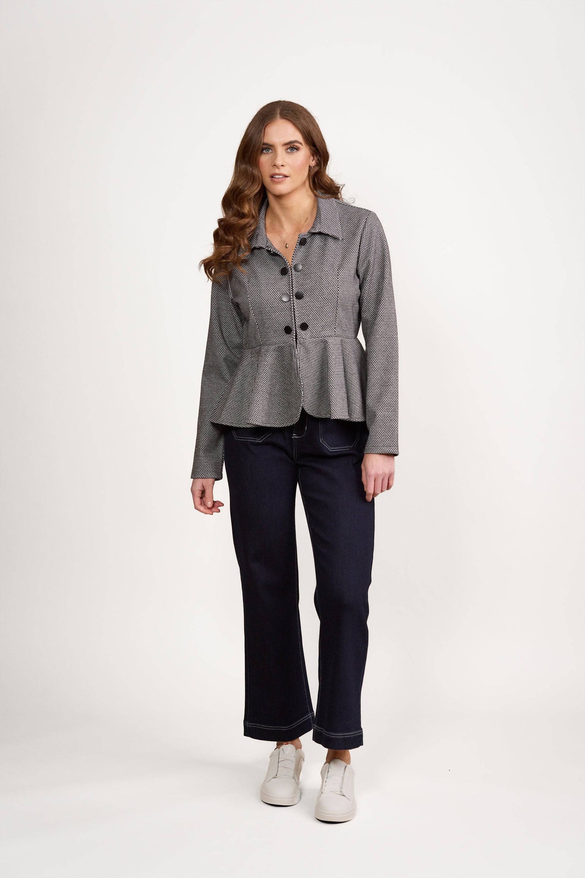 Woman with long wavy brown hair wearing the Vassalli Knit Jacket - Xander in gray, dark blue jeans, and white sneakers, standing against a plain white background.