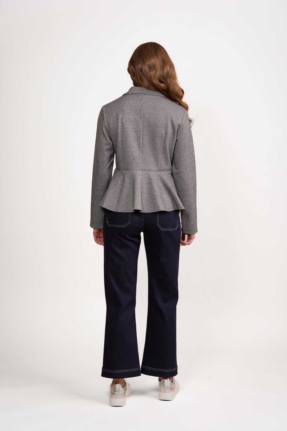 A person with long, wavy brown hair faces away, wearing Vassalli’s Knit Jacket - Xander in gray, paired with dark wide-leg jeans featuring white stitching and light platform shoes, against a white background.