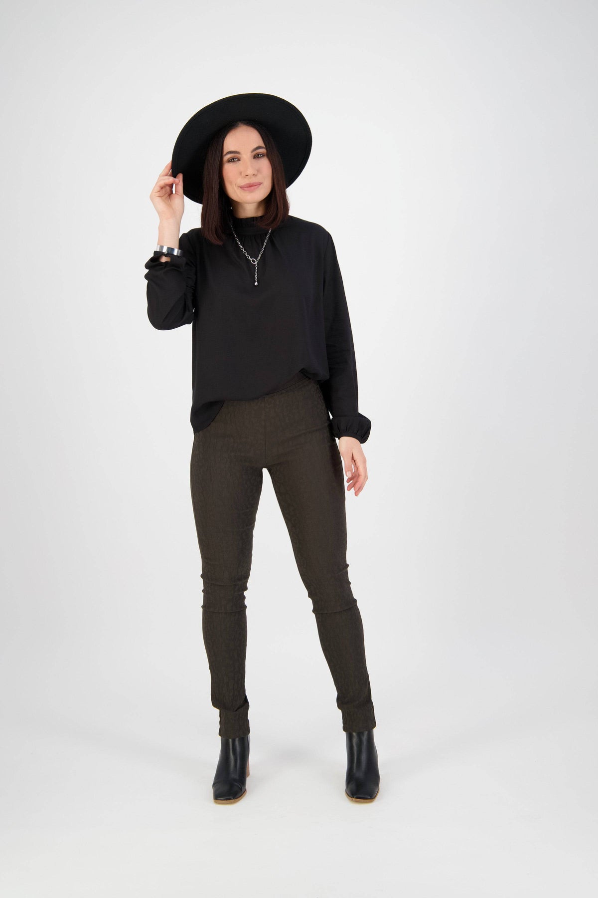A person in a black hat, black long-sleeve top, Vassalli Origin Pull On pants in olive, and black ankle boots stands against a plain white background, smiling and holding the brim of their hat.