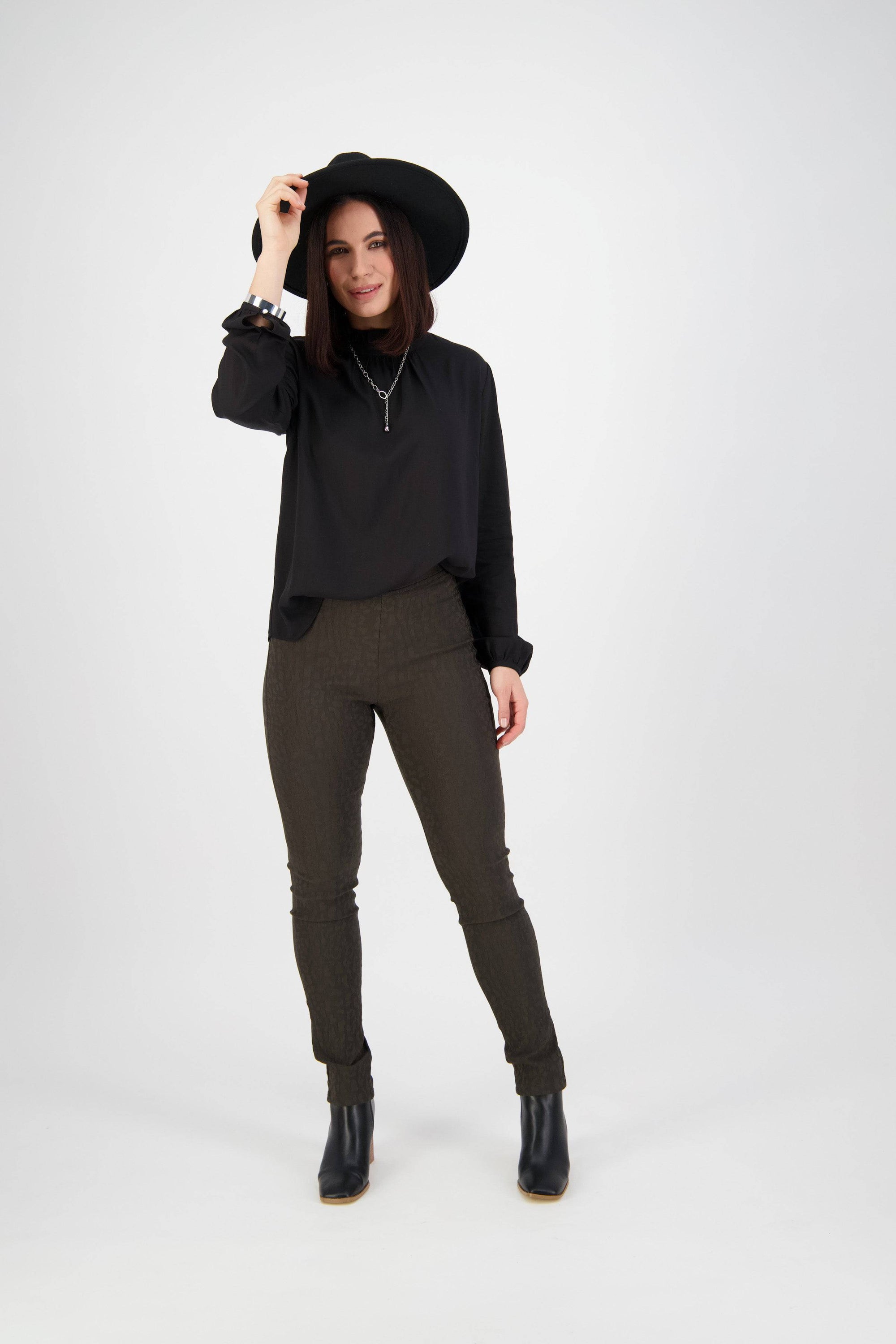 A woman stands against a white background, smiling and holding her black wide-brim hat. She wears a black long-sleeve top, Vassalli Origin Pull On - Olive pants, and black boots.