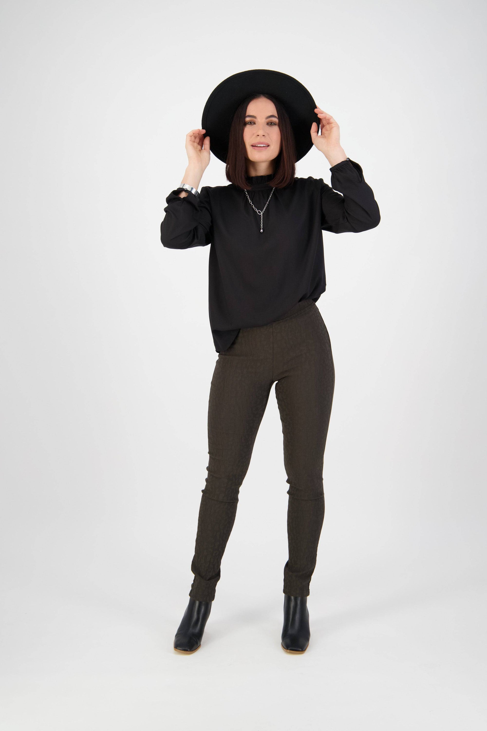 A person with straight dark hair wears a black wide-brim hat, black long-sleeve top, Vassalli Origin Pull On - Olive pants, and black ankle boots, standing against a plain white background and smiling slightly.