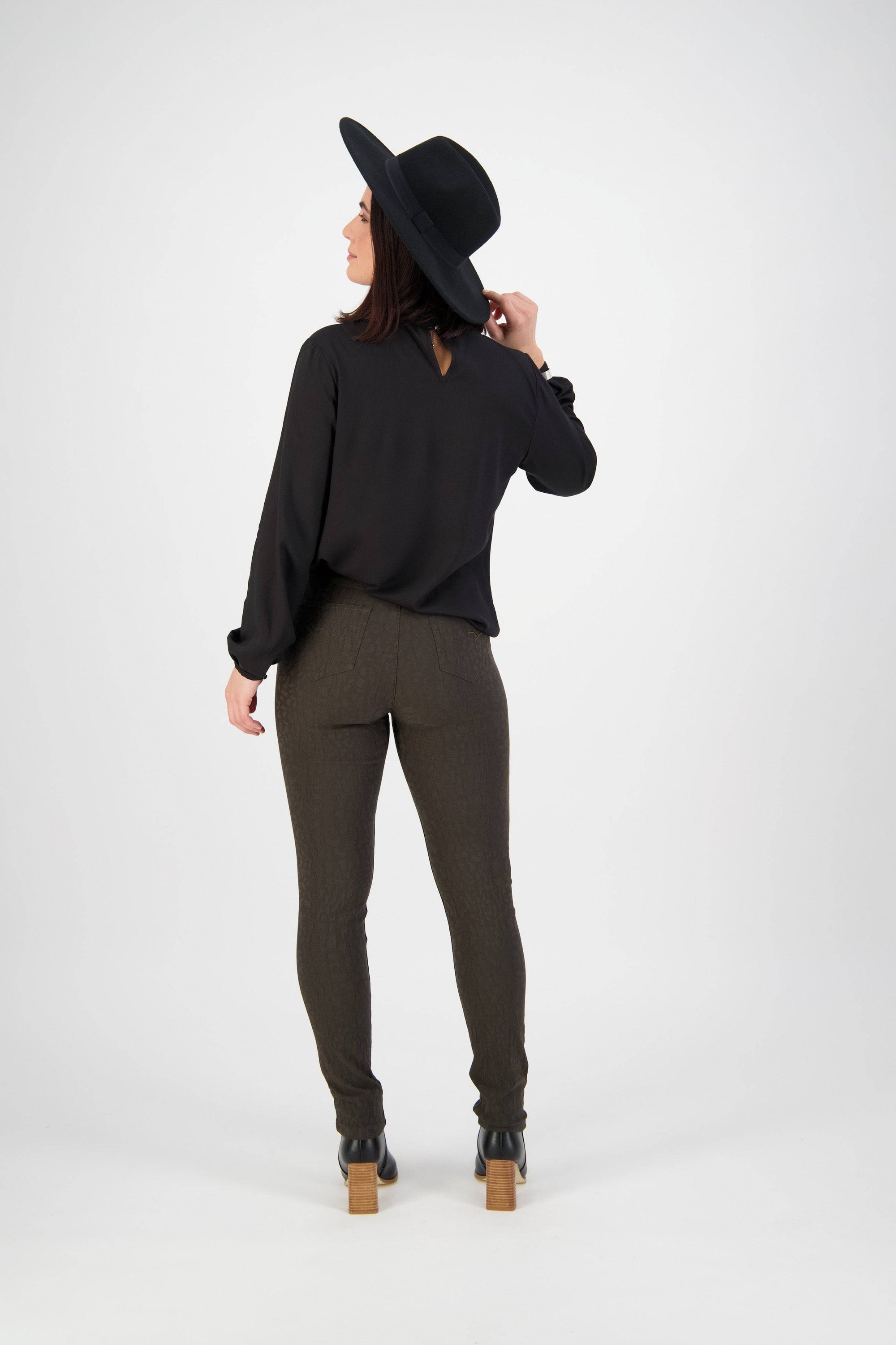 A woman stands facing away, wearing an olive Vassalli Origin Pull On pant, a black long-sleeve blouse, black heeled boots, and a wide-brimmed hat. She looks slightly left while holding the hat with one hand.