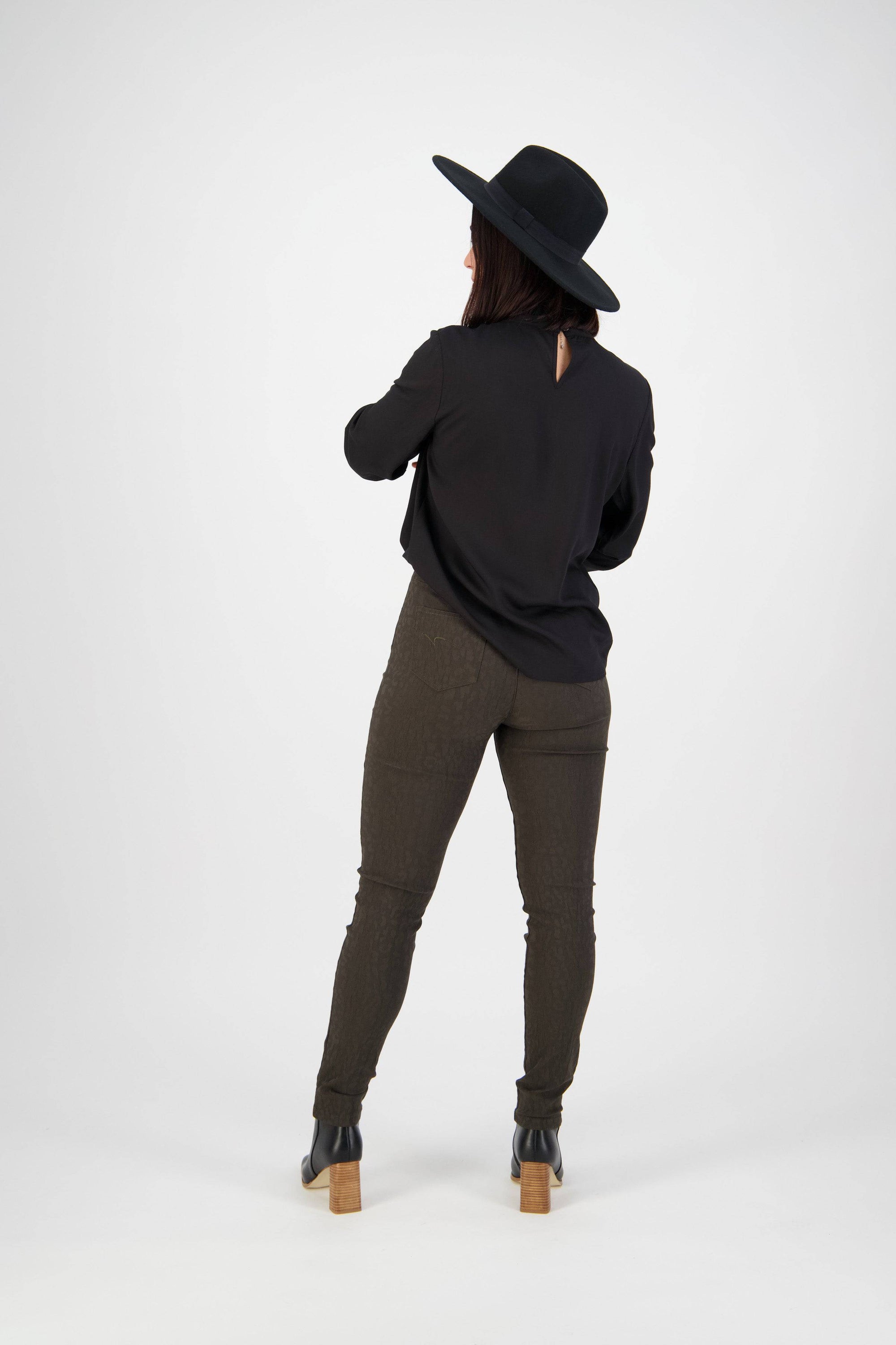 A person wearing a black wide-brim hat, black long-sleeve top, Vassalli Origin Pull On - Olive pants, and black ankle boots stands facing away against a plain white background.