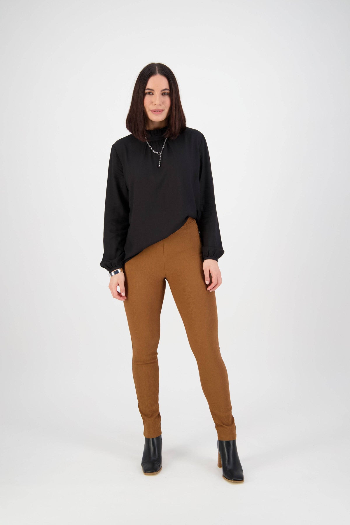 A woman with straight shoulder-length dark hair and a subtle smile faces the camera, wearing Vassalli Origin Pull On - Spice high rise skinny pants and a black long-sleeve top with black ankle boots against a plain white background.