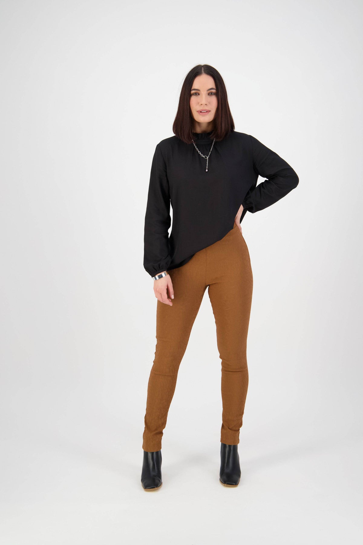 A woman with straight, shoulder-length dark hair stands against a white background wearing Vassalli's Origin Pull On - Spice pants, a black long-sleeve top, black boots, and layered necklaces.