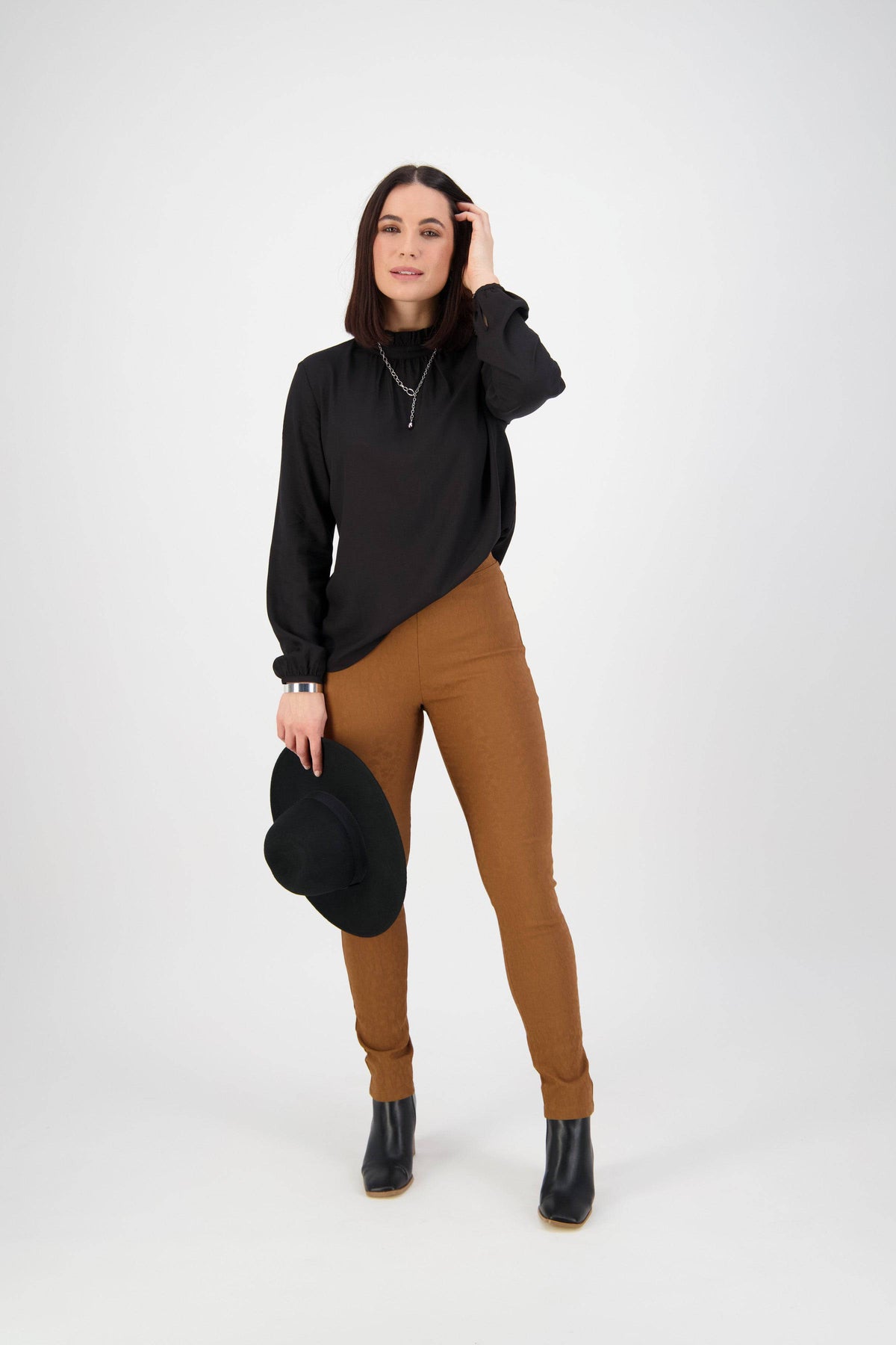 A woman stands against a white background in the Vassalli Origin Pull On - Spice pant, paired with a black long-sleeve top and black ankle boots. She holds a black hat in one hand, touching her hair with the other while gazing confidently at the camera.