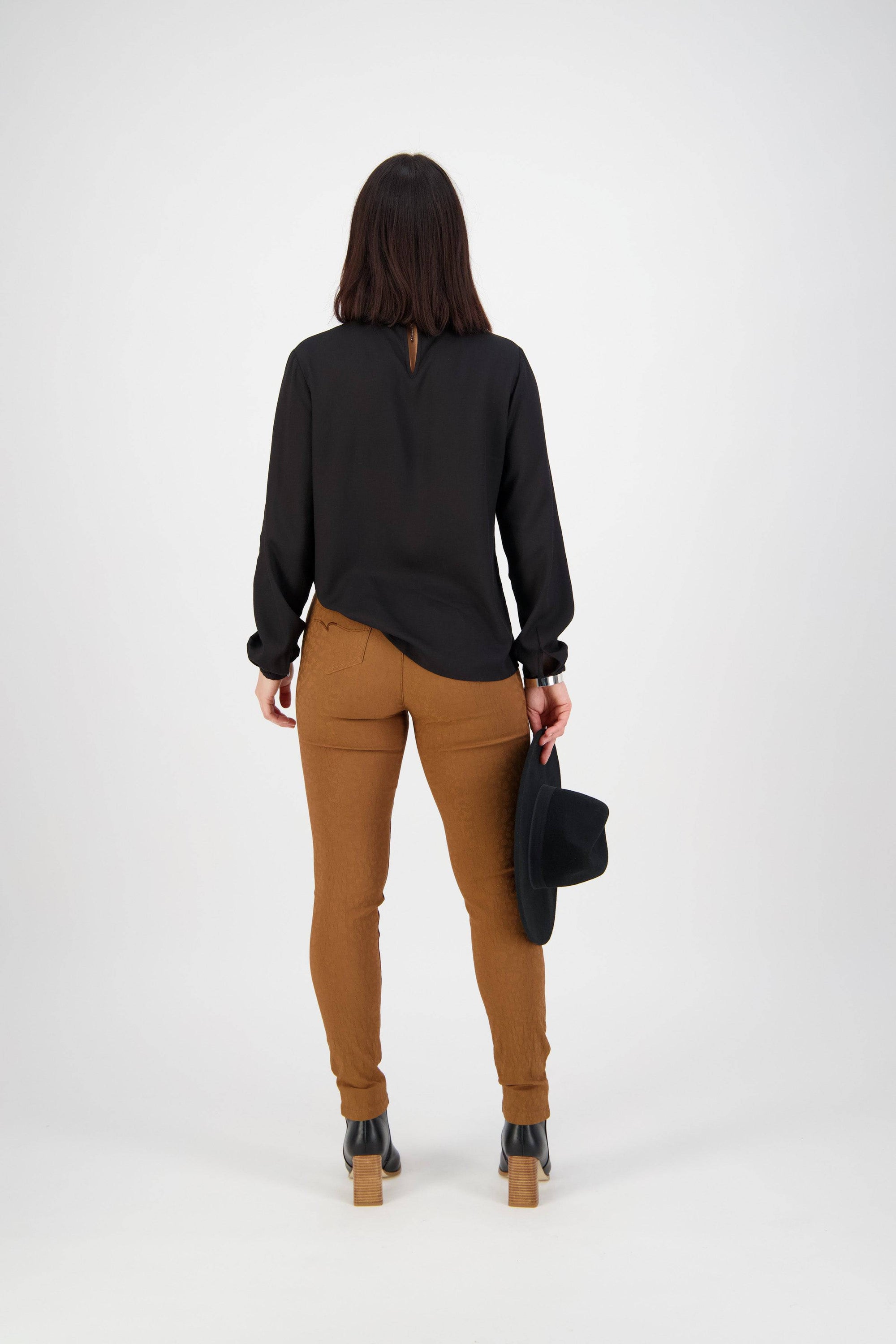 A person with long dark hair wears a black blouse, Vassalli Origin Pull On - Spice high rise brown skinny pants, and black heeled boots, standing against a light background holding a black hat in their right hand.