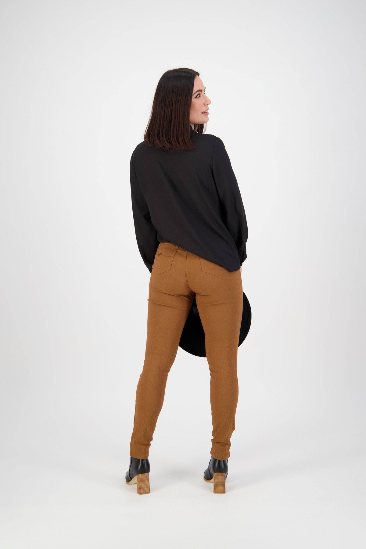 A woman with straight brown hair stands facing away, wearing Vassalli’s Origin Pull On - Spice high rise, skinny leg pants and a black blouse, paired with black heeled boots and holding a black hat against a white background.