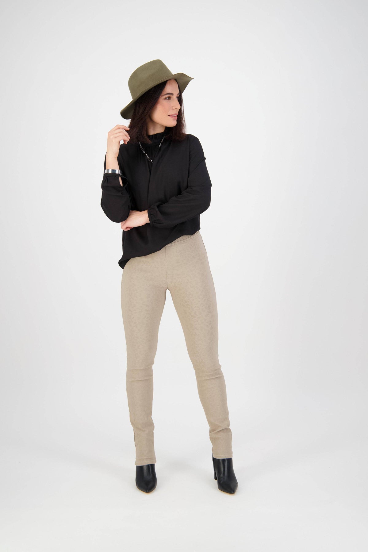 A woman in a green hat, black long-sleeve top, Vassalli Origin Pull On - Taupe pants, and black boots stands against a white background, looking to the side with one hand touching her hat.
