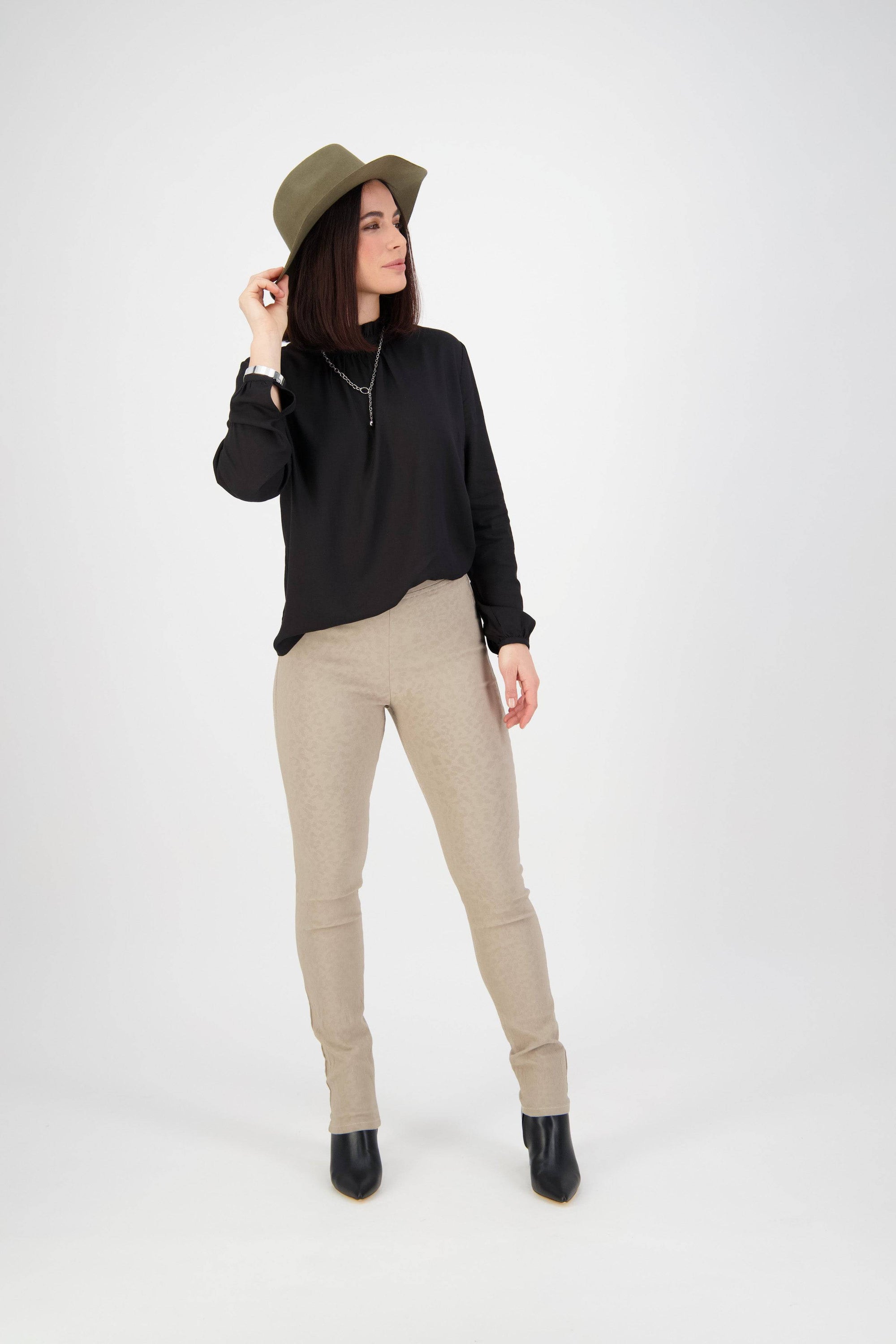 A woman stands against a white background wearing a wide-brim hat, black long-sleeve top, Vassalli Origin Pull On pants in taupe, and black ankle boots, looking to the side while holding her hat with one hand.