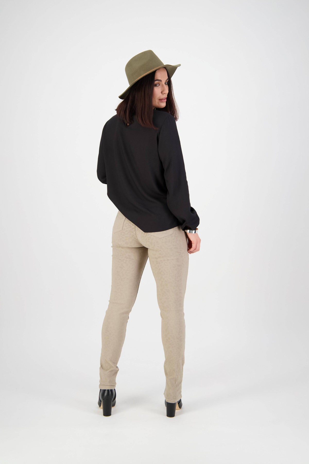A woman stands looking over her shoulder, wearing a green hat, black long-sleeve top, Vassalli Origin Pull On - Taupe pants, and black heeled boots against a plain white background.