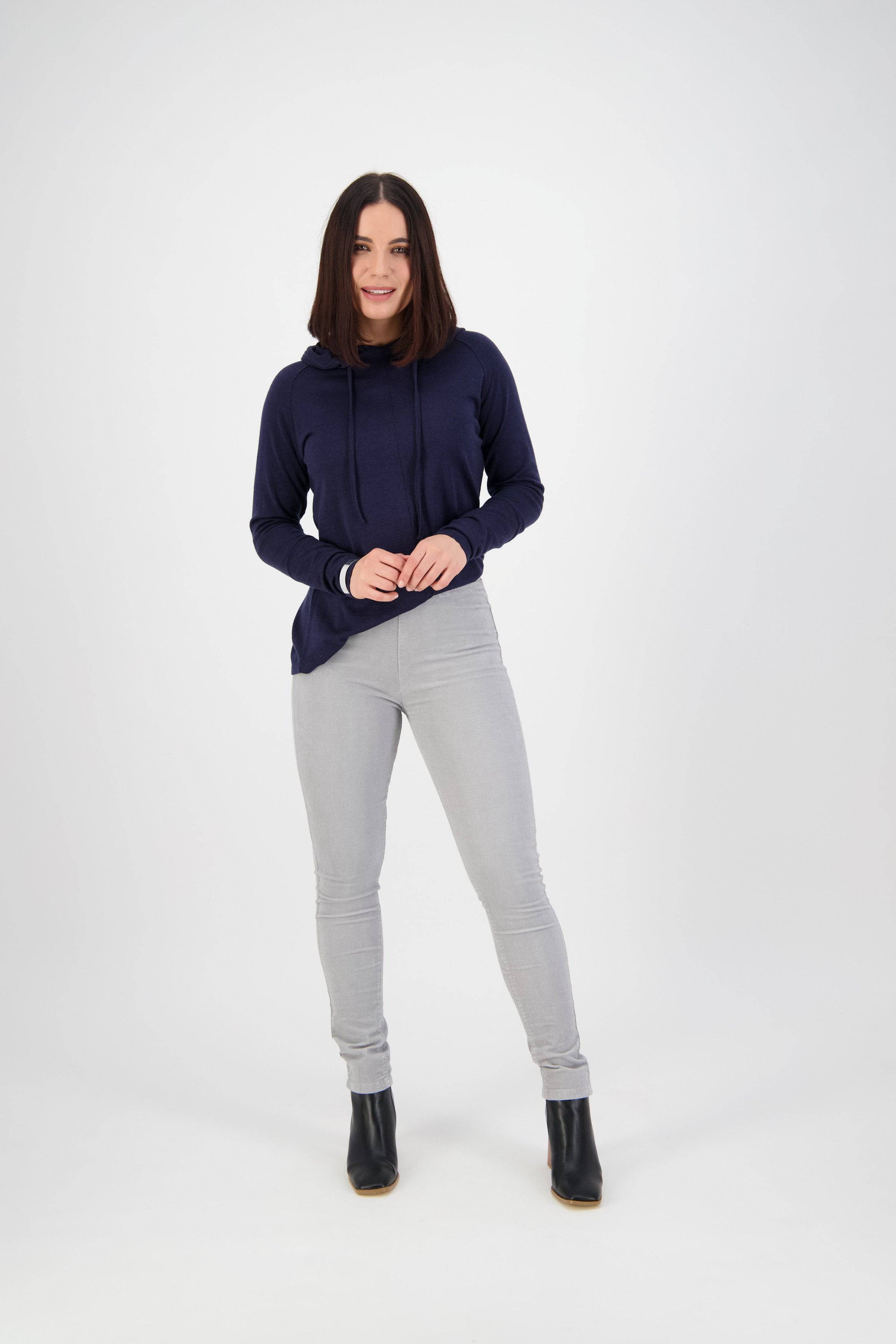 A woman with straight brown hair wears a navy blue hoodie, Vassalli Origin Cord Pull On in Cloud, and black ankle boots as she smiles against a plain white background.
