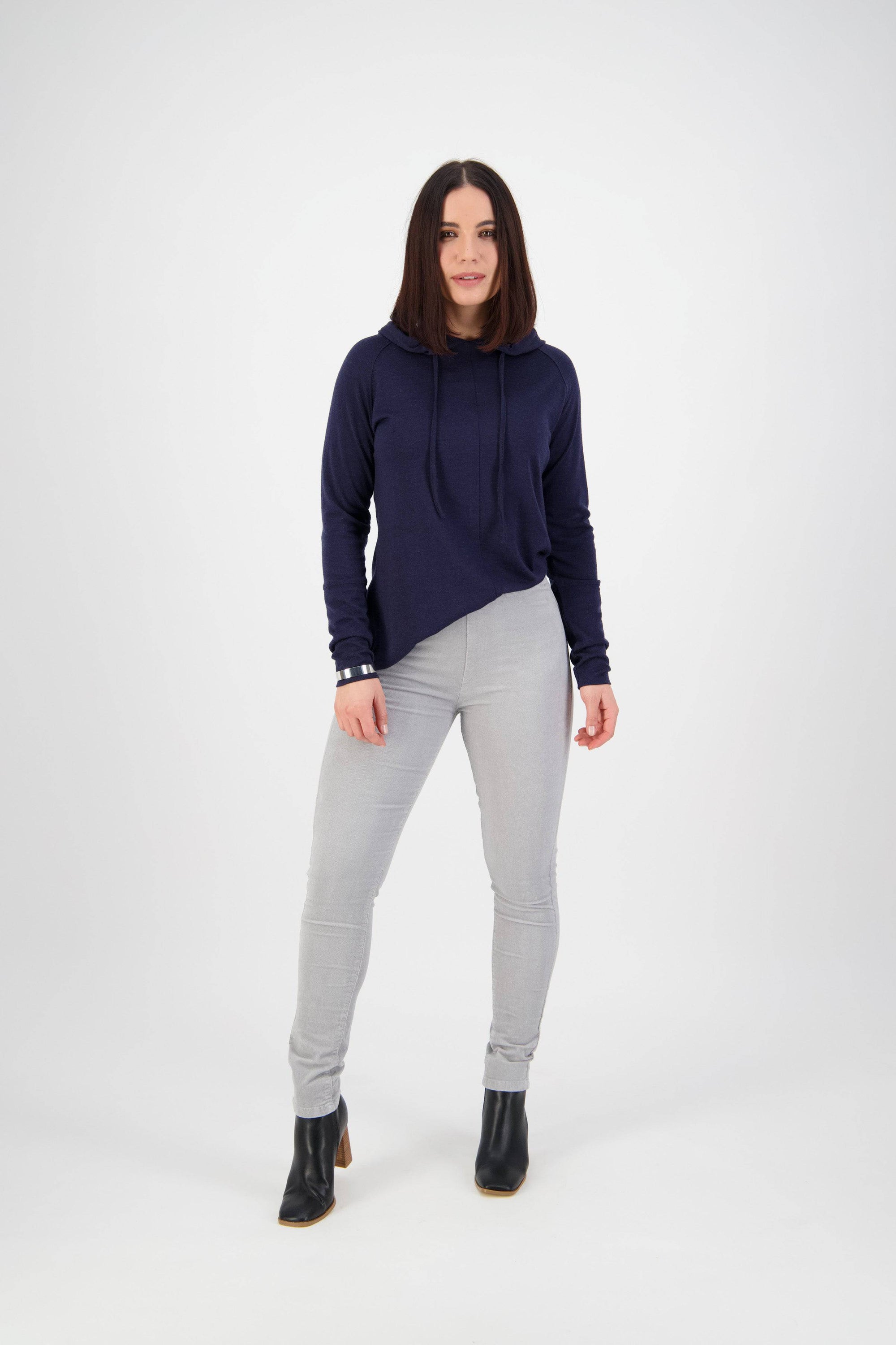 A woman with straight dark hair faces forward, wearing a navy hoodie, Vassalli Origin Cord Pull On pants in Cloud, and black ankle boots against a plain white background.