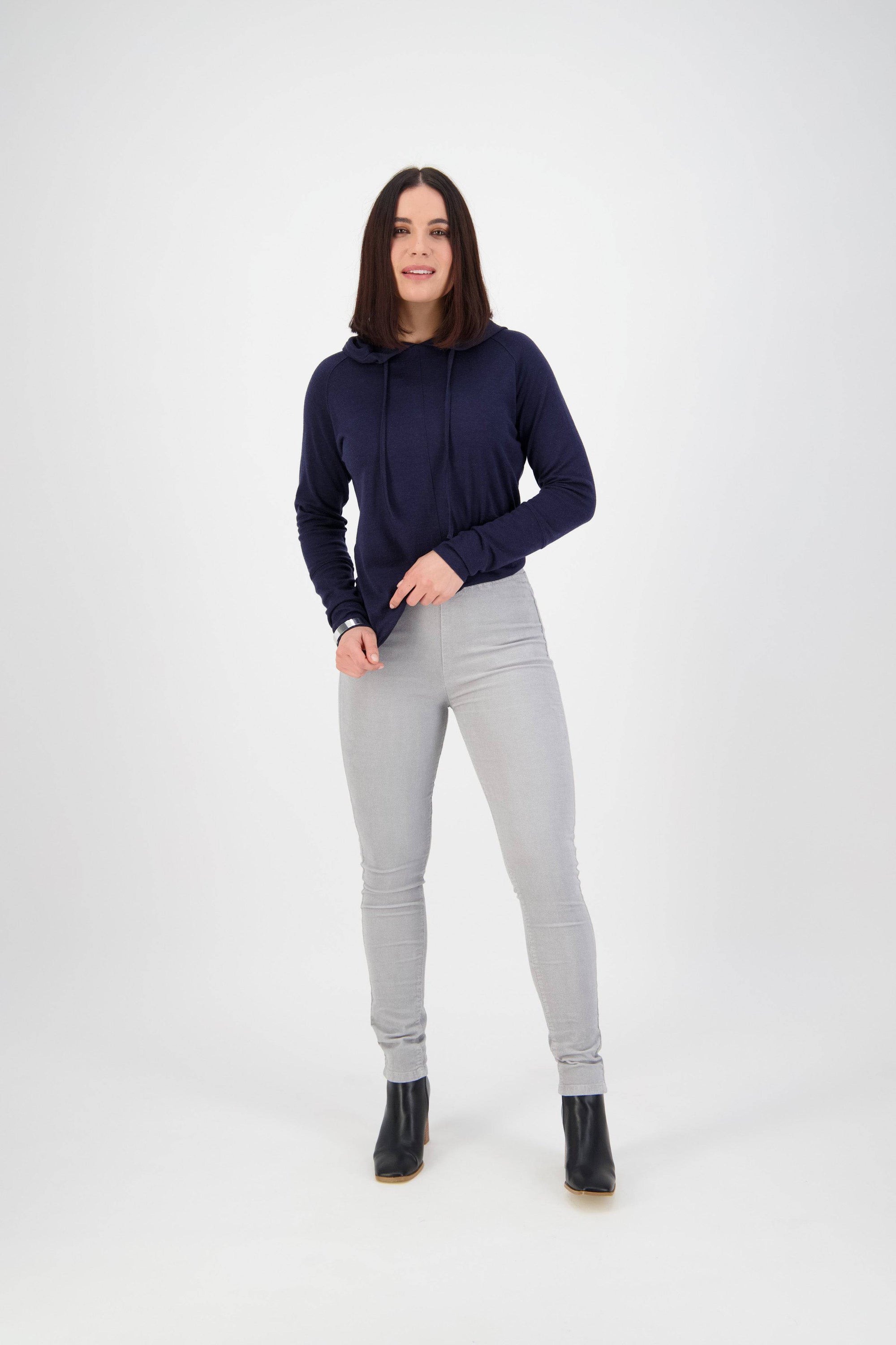 A woman with straight, shoulder-length dark hair stands against a white background, wearing a navy blue hoodie, Vassalli Origin Cord Pull On pants in Cloud, and black ankle boots. She looks forward and smiles slightly.