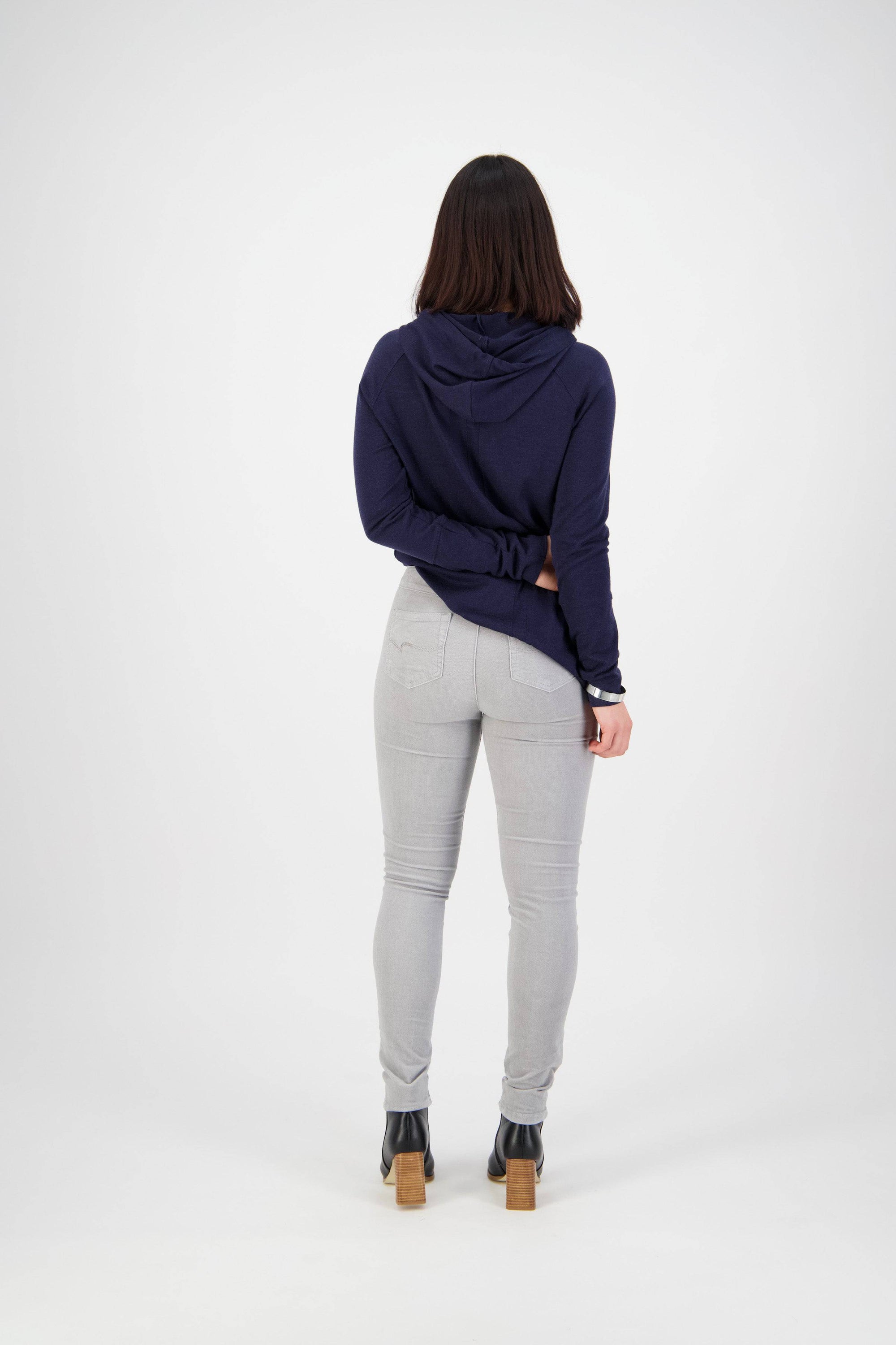 A person with shoulder-length dark hair, wearing a navy hoodie, Vassalli Origin Cord Pull On pants in Cloud, and black heeled boots stands facing away from the camera against a plain white background.