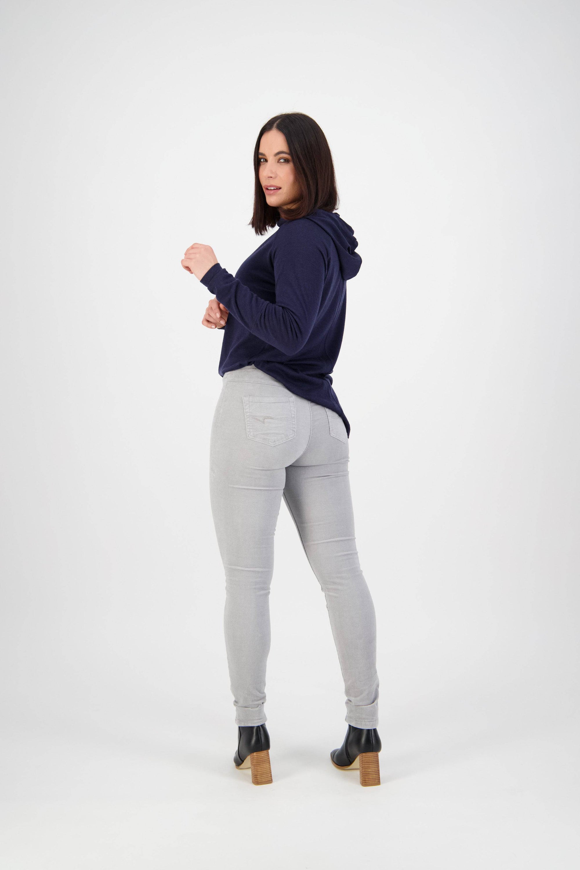 A woman with shoulder-length dark hair stands in a studio, wearing a navy hoodie, Vassalli Origin Cord Pull On pants in Cloud, and heeled ankle boots, looking over her shoulder toward the camera.