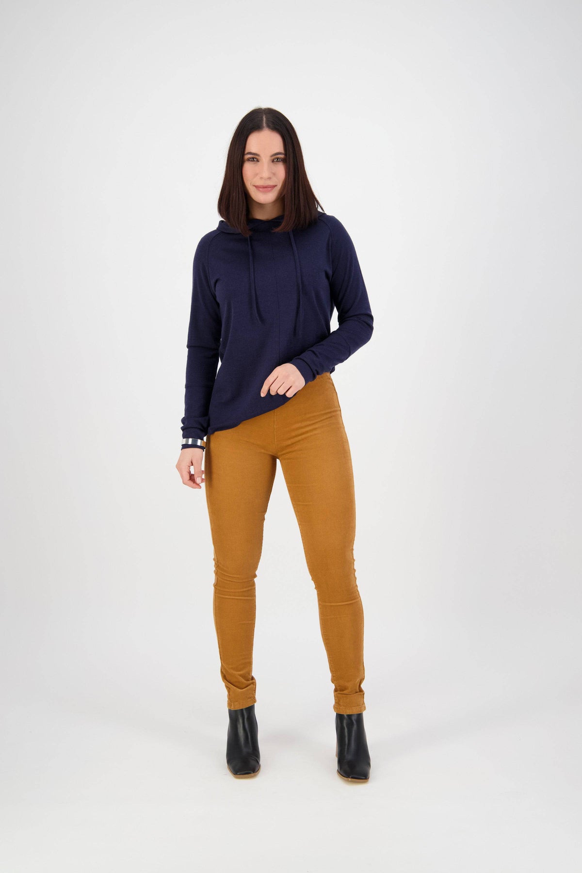 A woman with straight dark hair stands against a white background, wearing a navy blue hoodie and Vassalli Origin Cord Pull On pants in Copper, paired with black ankle boots. She faces forward with one hand in her pocket and smiles slightly.