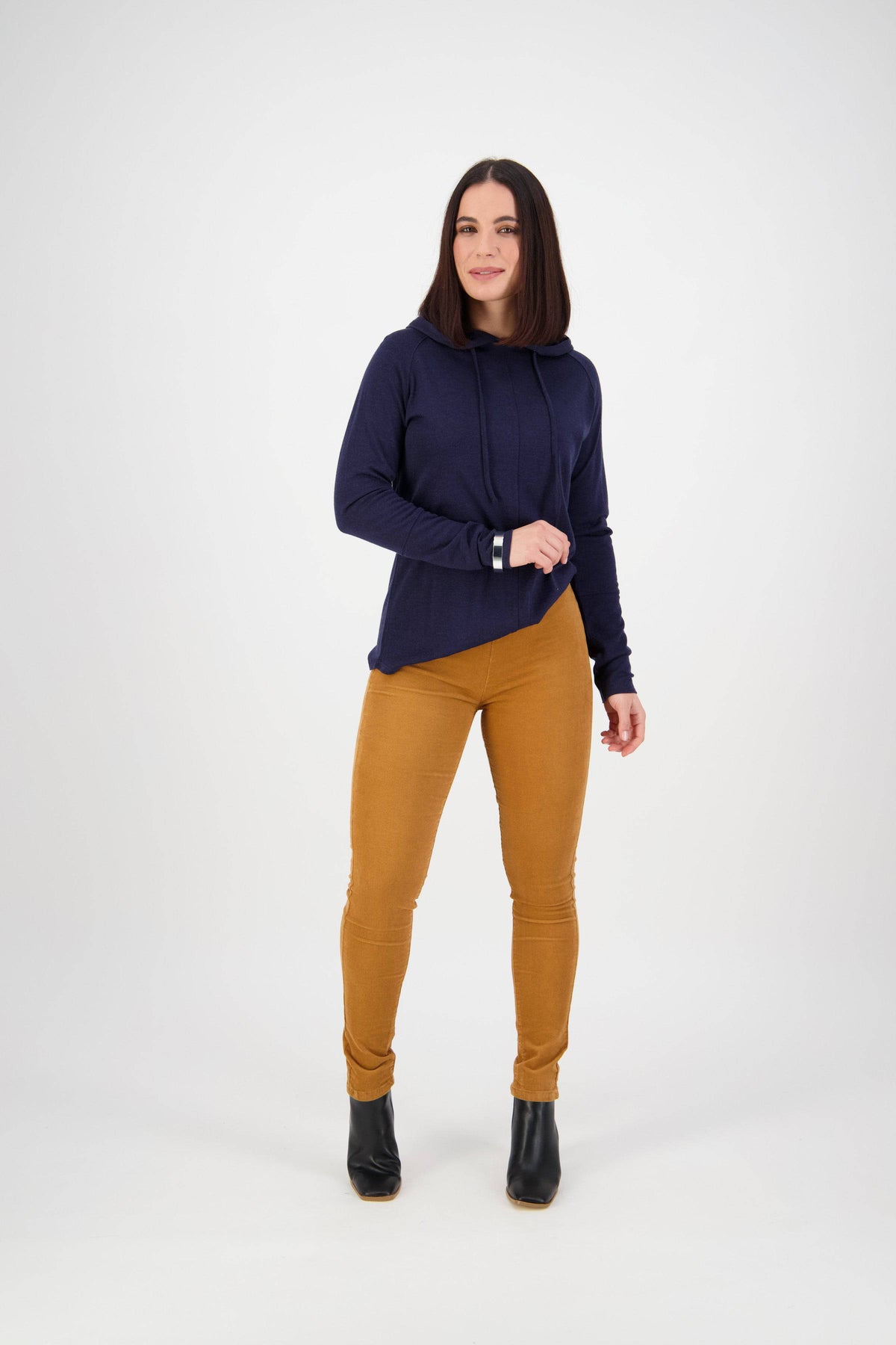 A woman with straight dark hair stands against a white background, wearing a navy hoodie, Vassalli Origin Cord Pull On pants in Copper, and black ankle boots. She faces forward with a neutral expression.