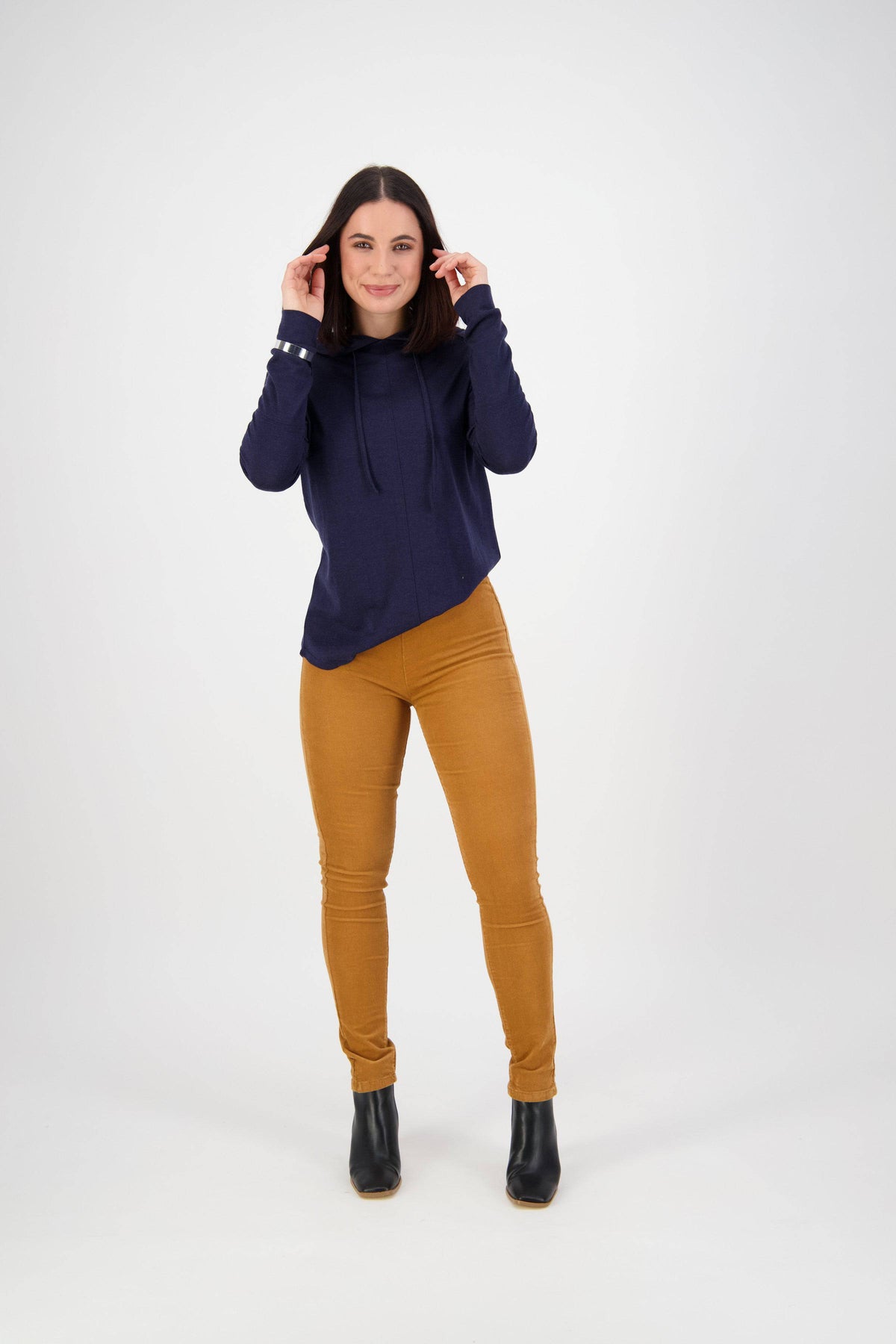 A woman smiles and touches her hair while wearing a navy blue hoodie, black ankle boots, and Vassalli’s Origin Cord Pull On pants in Copper, standing against a plain white background.