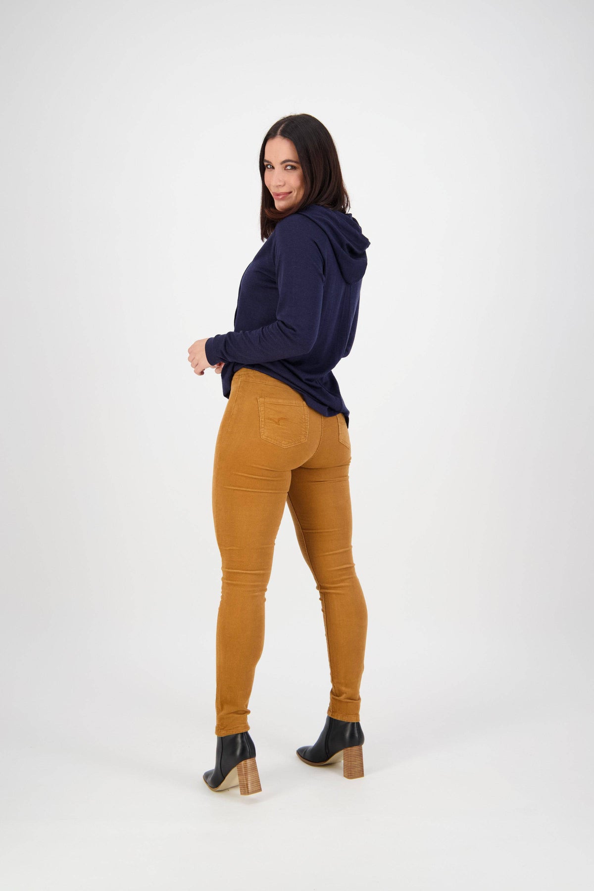 Woman stands against a plain white background, looking over her shoulder in a navy blue hoodie, Vassalli Origin Cord Pull On - Copper high rise skinny pants, and black ankle boots with wooden heels.