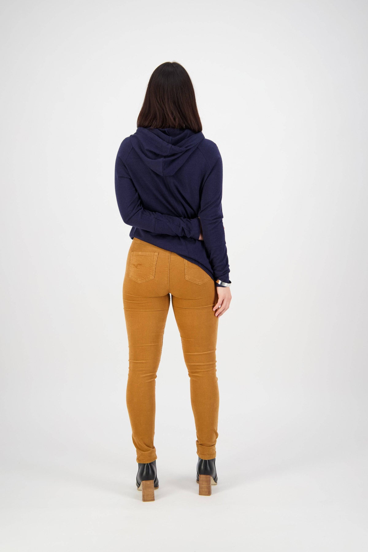 A woman faces away from the camera, wearing a navy blue hoodie, black heeled boots, and Vassalli's Origin Cord Pull On pants in Copper, set against a plain white background.