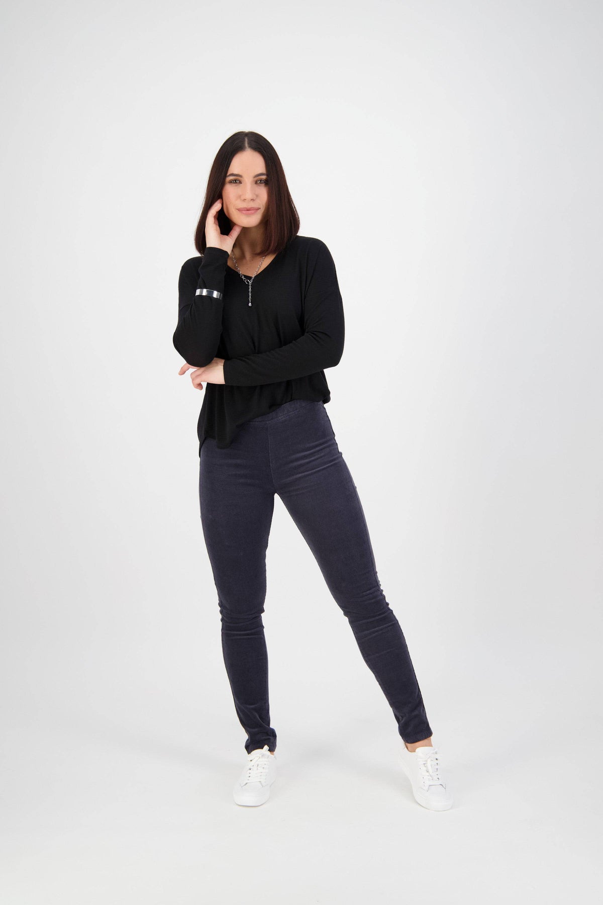 A woman with straight dark hair poses against a white background, wearing the Vassalli Origin Cord Pull On - Graphite pants, a black long-sleeve top, and white sneakers. She rests her chin on her hand and smiles softly.