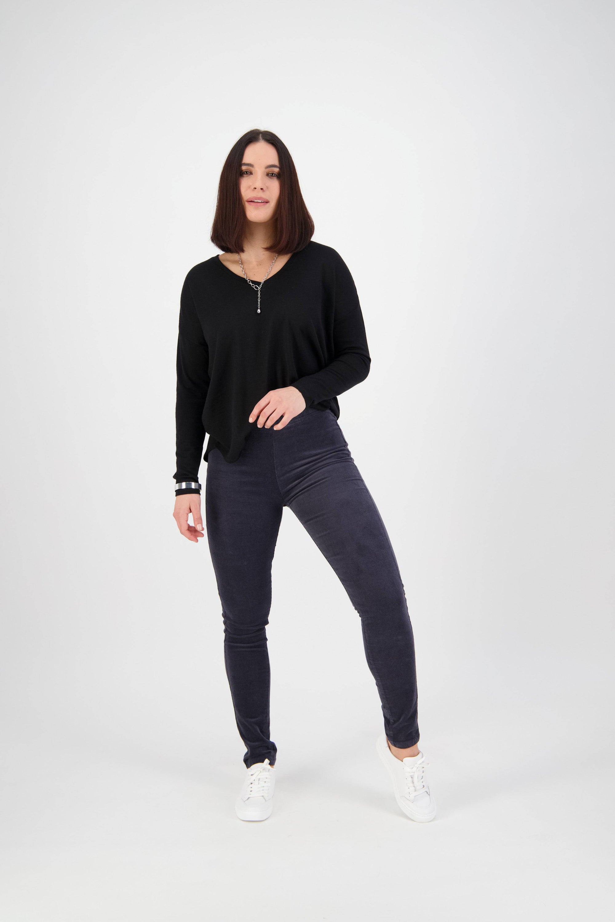 A woman with shoulder-length brown hair stands against a white background, wearing the Vassalli Origin Cord Pull On pants in Graphite and a black long-sleeve top with white sneakers, looking at the camera with a relaxed expression.