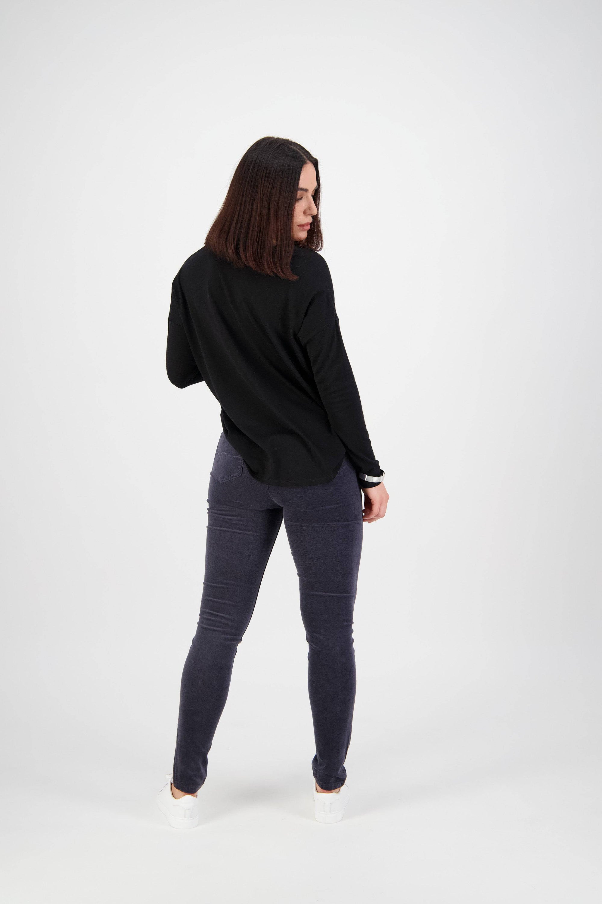A woman with straight, shoulder-length brown hair faces away, turning slightly right. She wears a black long-sleeve top and Vassalli Origin Cord Pull On - Graphite slim leg pants with white sneakers against a plain white background.