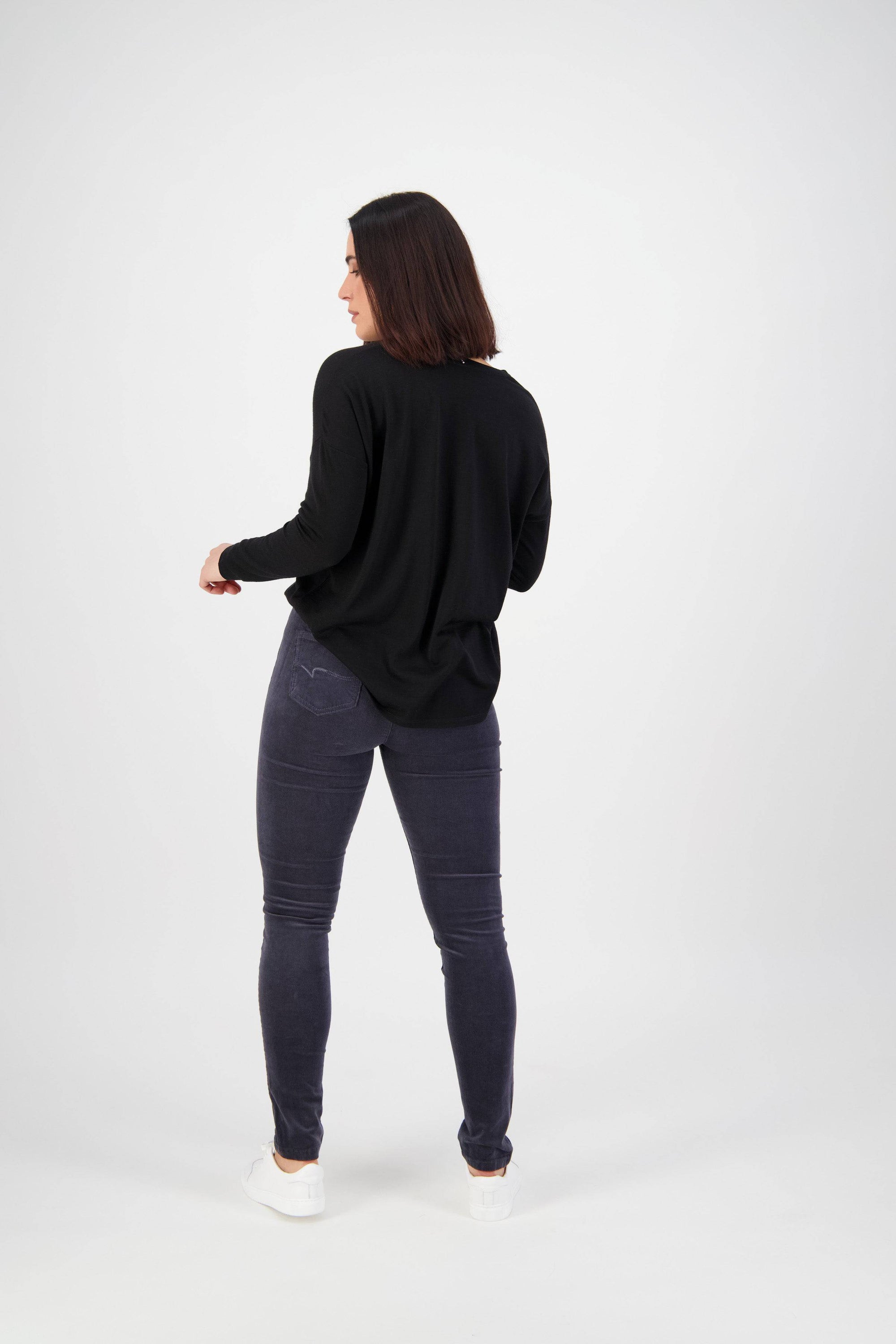 A person with shoulder-length dark hair, wearing a black long-sleeve top, Vassalli Origin Cord Pull On - Graphite slim leg pants, and white shoes stands facing away from the camera against a plain white background.