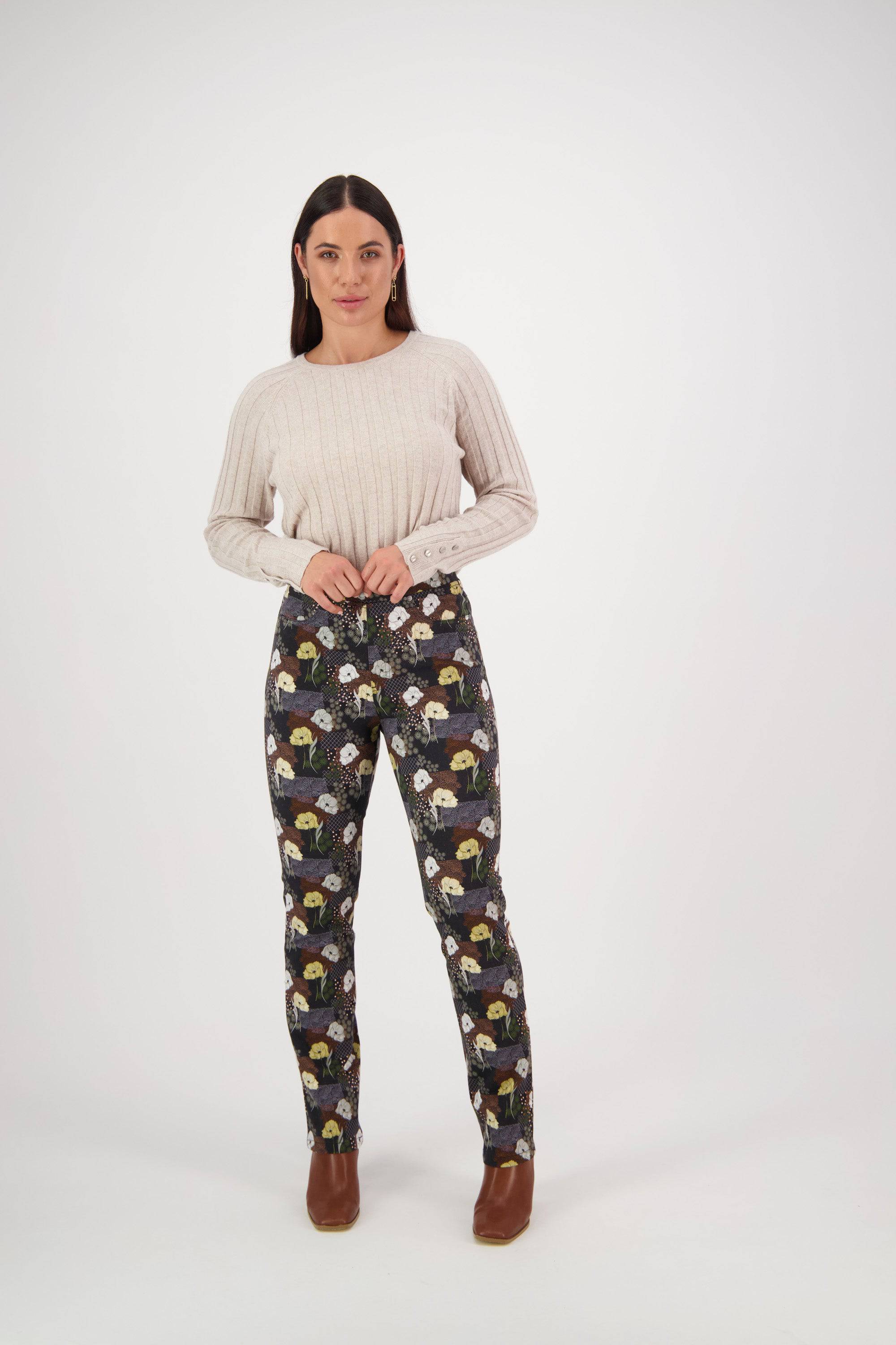 A woman stands facing forward against a plain background, wearing a cream ribbed sweater and Vassalli's Bay Pull On - Karma slim leg pants in a colorful floral print, made from stretch fabric. Her hands are at her waist and she wears brown shoes.