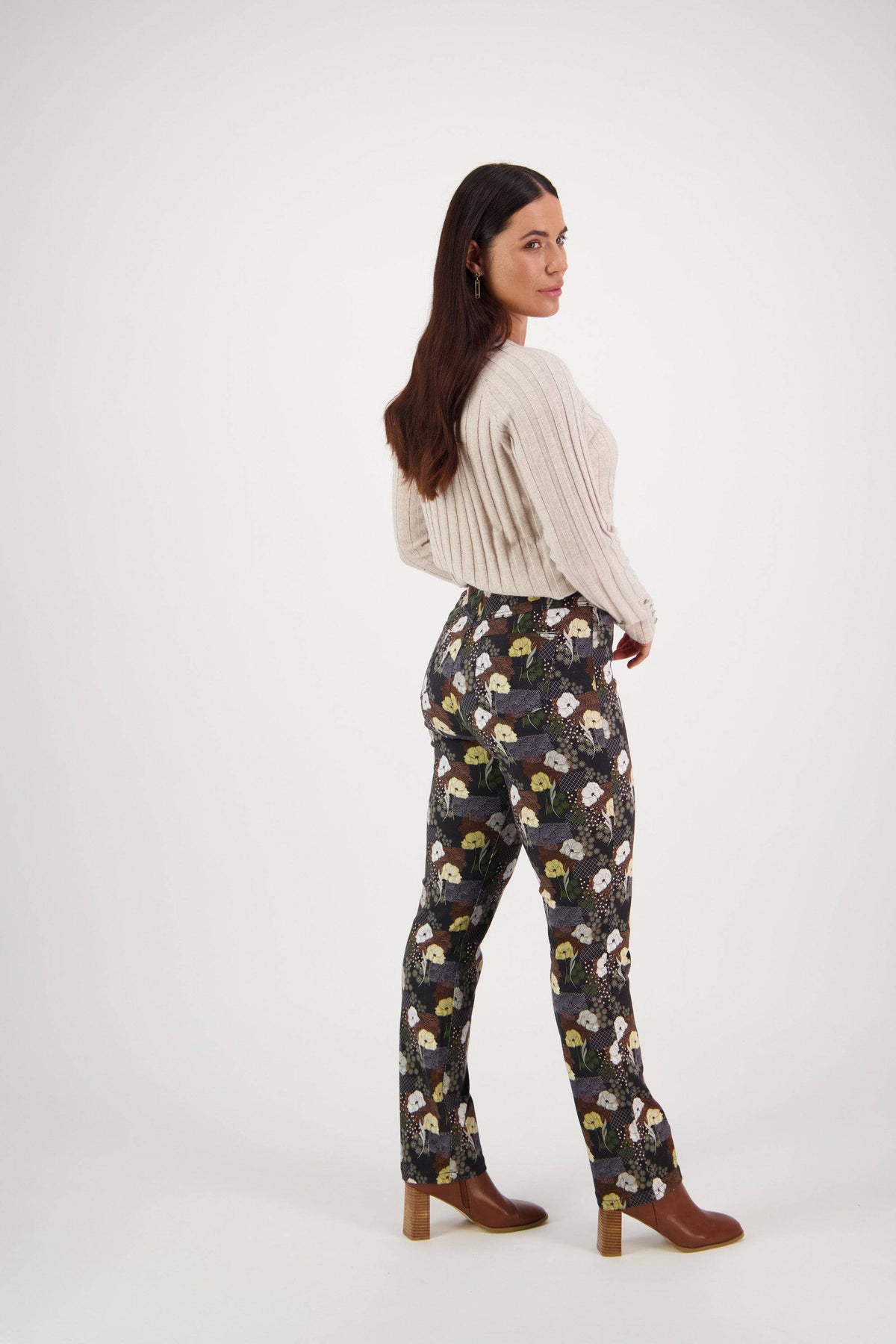 A woman with long dark hair wears a cream cable-knit sweater, Vassalli Bay Pull On - Karma slim leg printed pants, and brown heeled boots, standing sideways and looking over her shoulder against a plain white background.