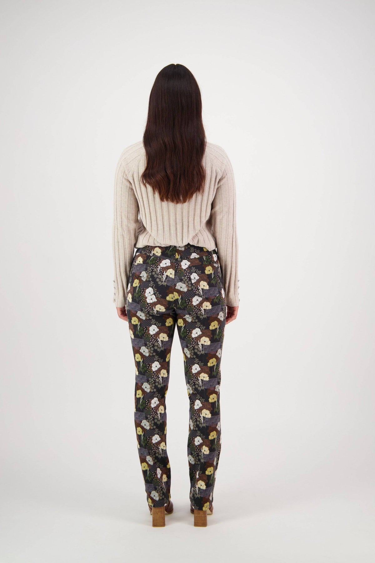 A person with loose dark hair poses against a white background, wearing a beige ribbed sweater and Vassalli's Bay Pull On - Karma slim leg pants, which feature a colorful dog print.