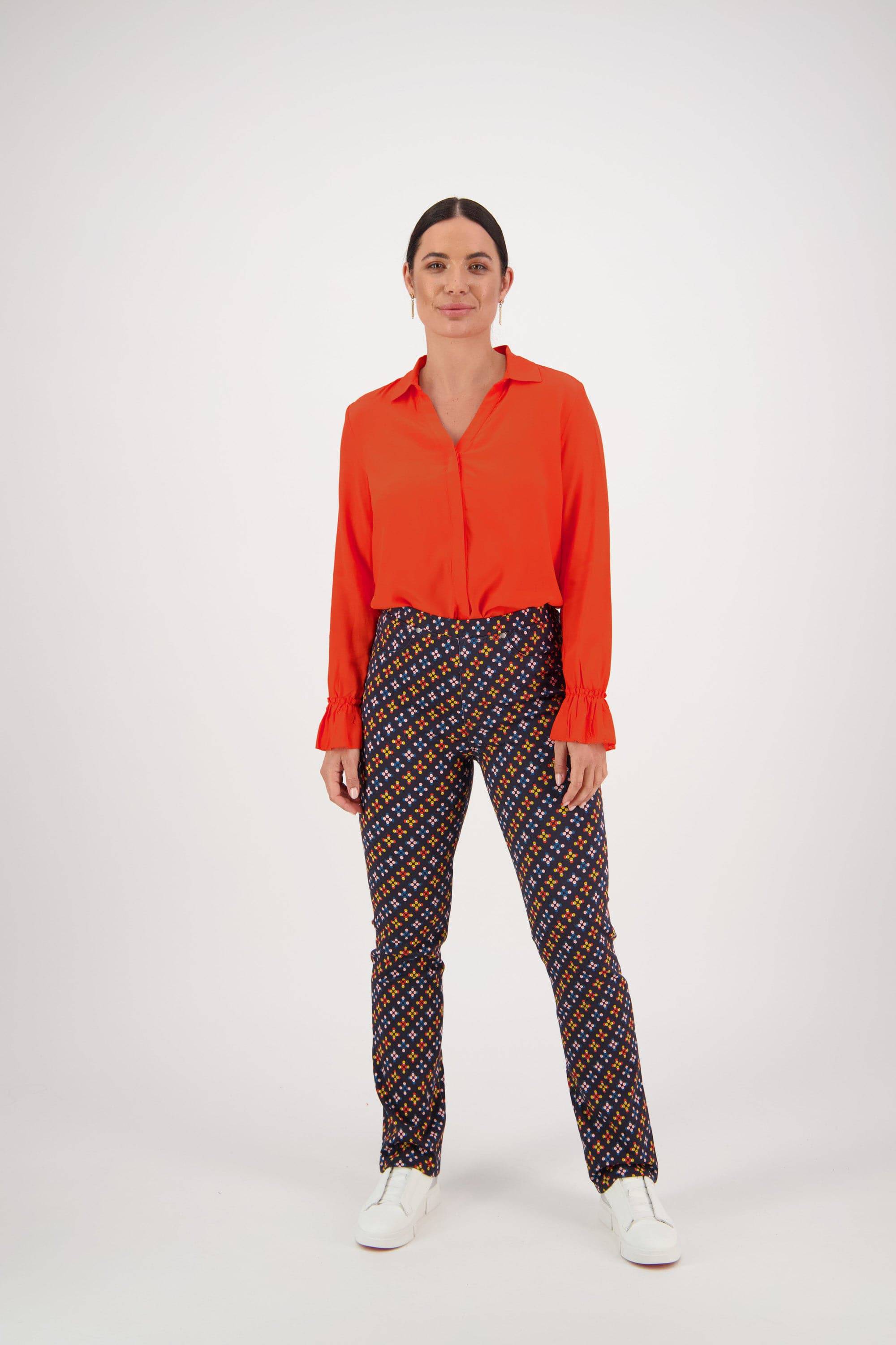 A woman faces forward against a white background, wearing a bright orange long-sleeve blouse, Vassalli Bay Pull On - Stellar slim leg pants, and white sneakers.