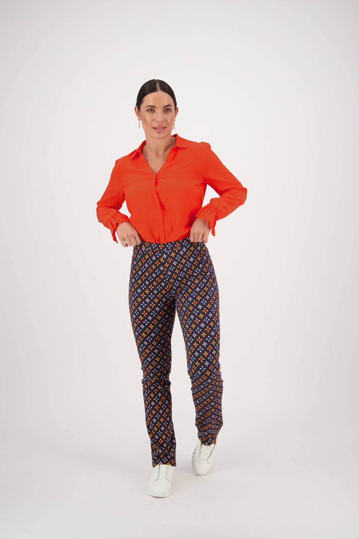 A woman stands against a white background wearing a bright red blouse, Vassalli Bay Pull On - Stellar slim leg pants, and white sneakers. She looks forward with her hands in her pockets.